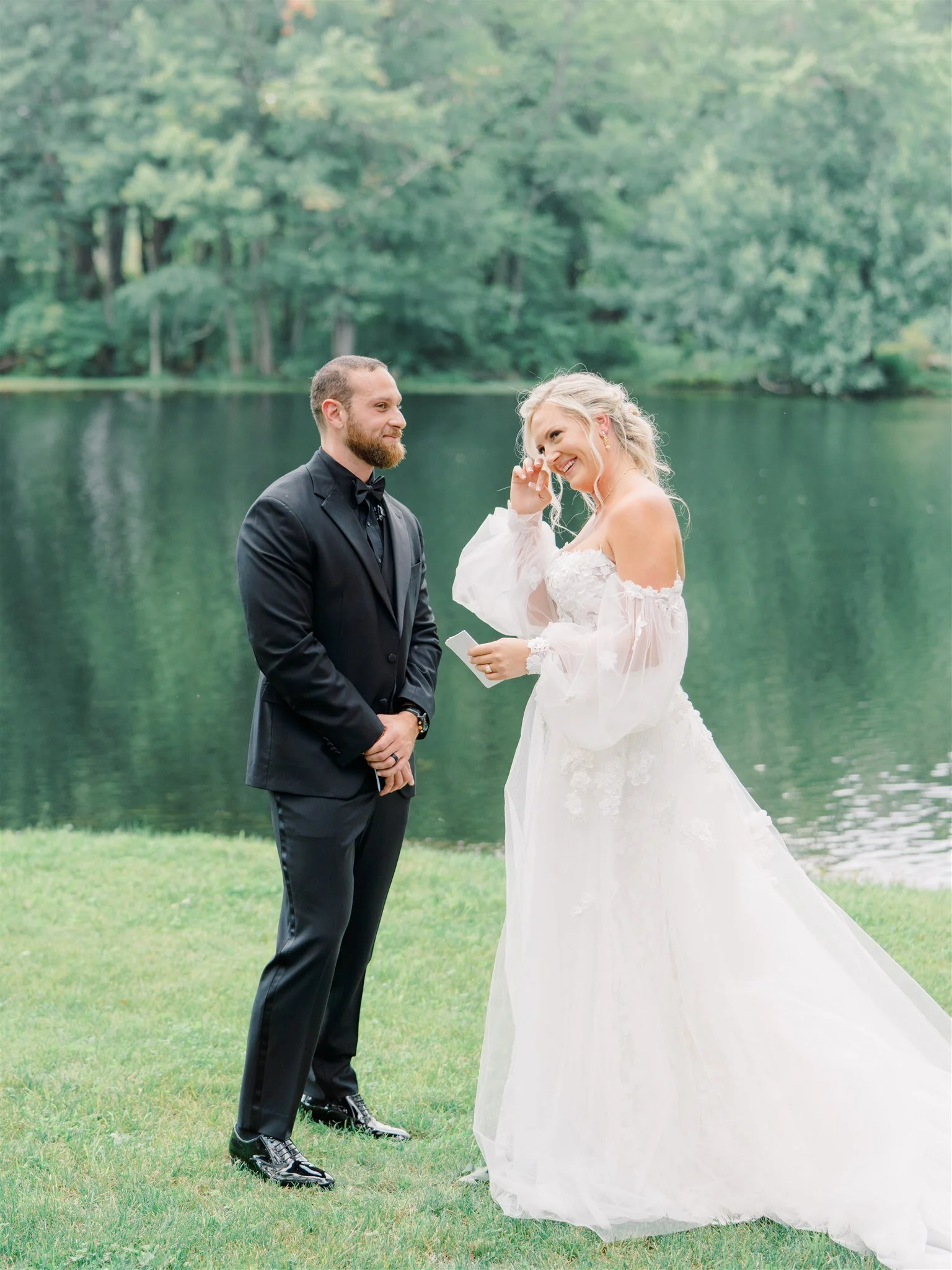 A bride and groom exchanging vows outdoors near a lake, with trees in the background.
