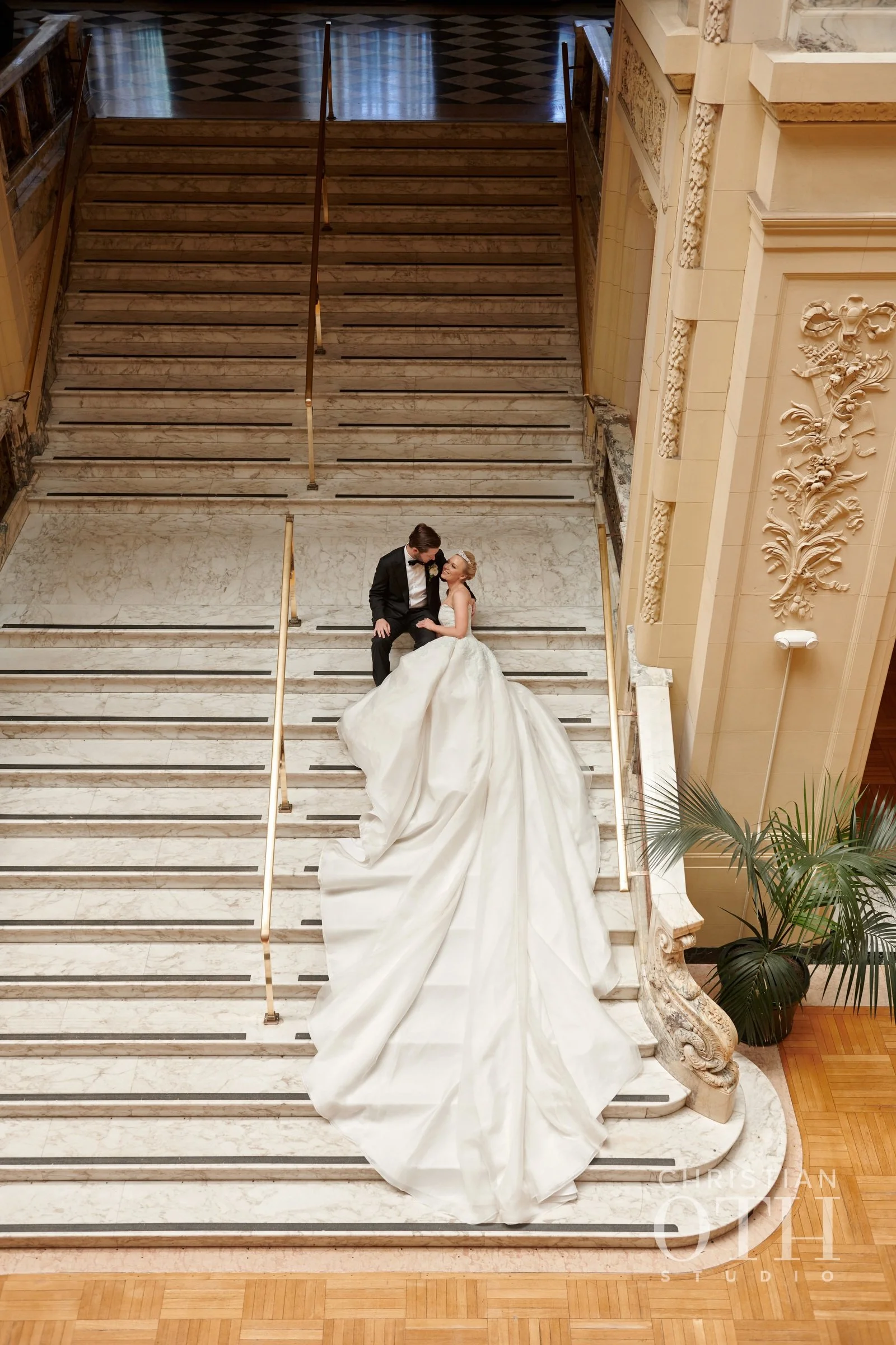 A bride and groom sitting on a wide marble staircase in a grand, ornate building. The bride is wearing a long, white wedding dress with a train, and the groom is in a black tuxedo. They are gazing at each other affectionately.
