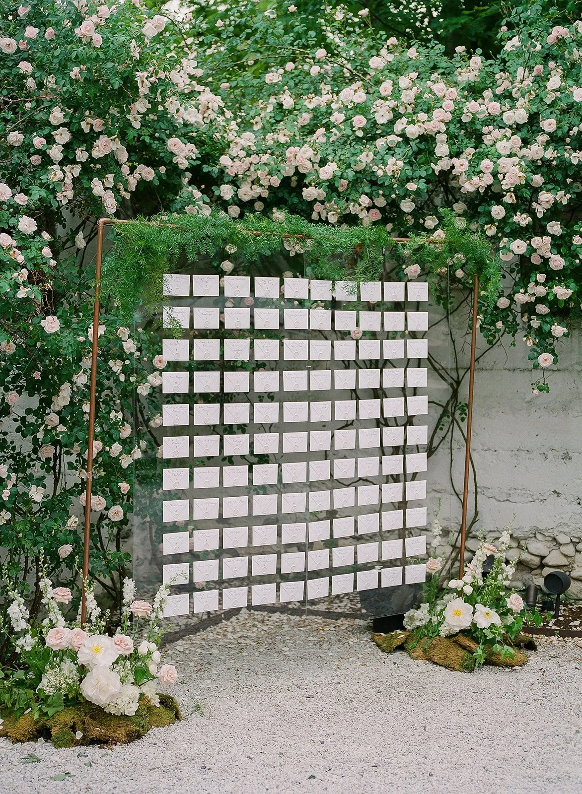 A rustic wedding or event seating chart with white cards pinned to a black grid, decorated with greenery and pink and white flowers.