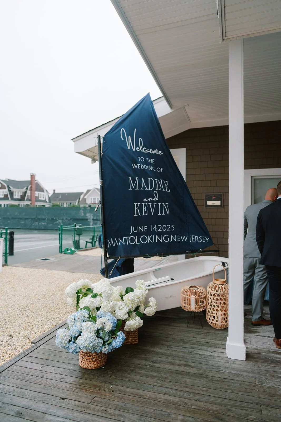 Wedding welcome sign with white text on a navy sailboat, decorated with white and blue hydrangeas and wicker lanterns, set on a wooden deck outside a house, with people dressed in formal suits in the background.