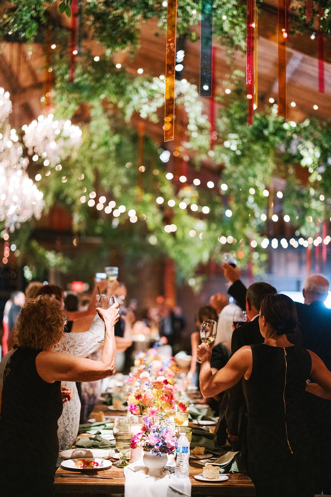 People at a wedding or formal event raising glasses for a toast at a decorated dinner table in a festive, warmly lit venue with greenery and hanging ribbons.