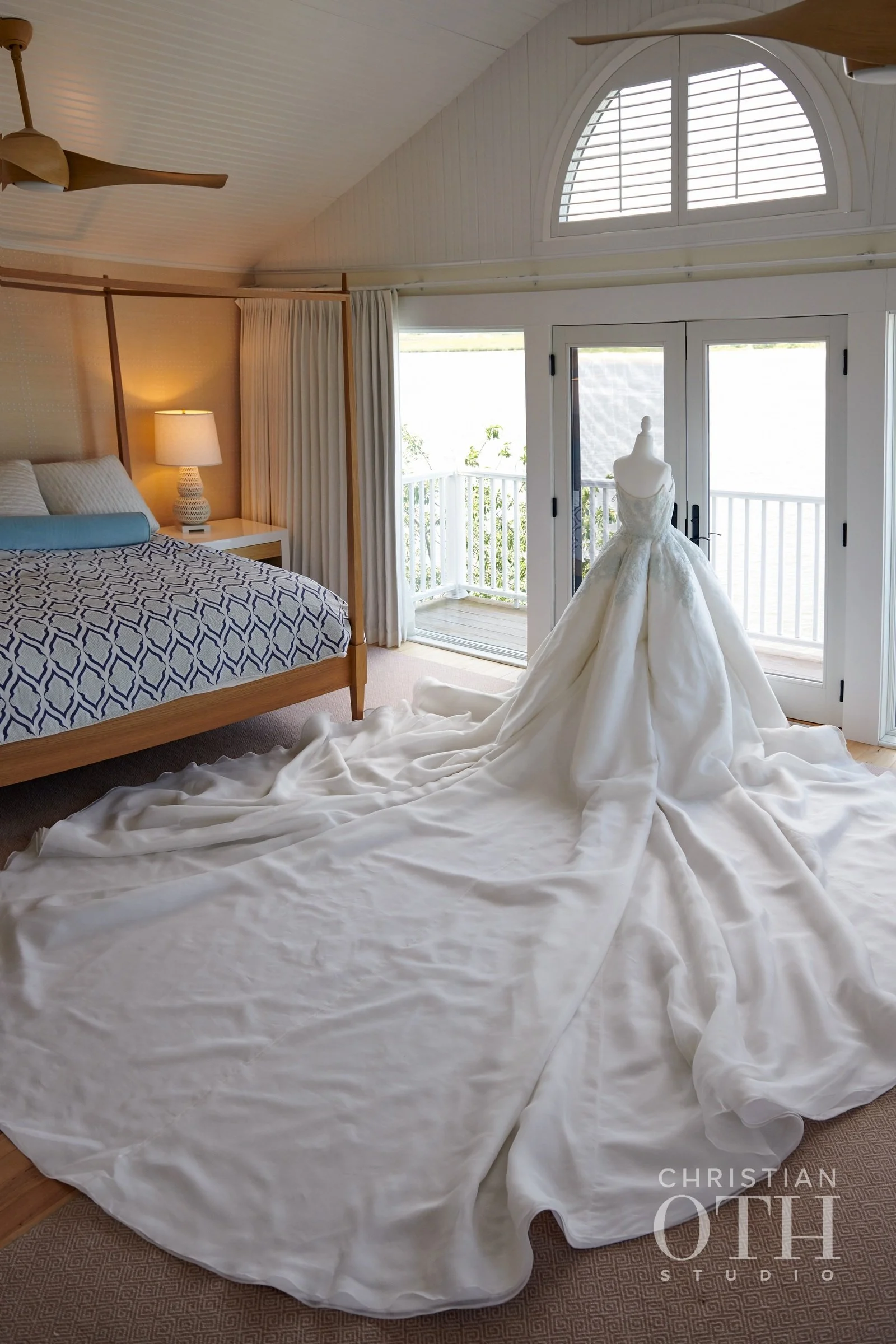 A wedding dress on a mannequin positioned on a bed in a bright bedroom with a sliding glass door and balcony outside.