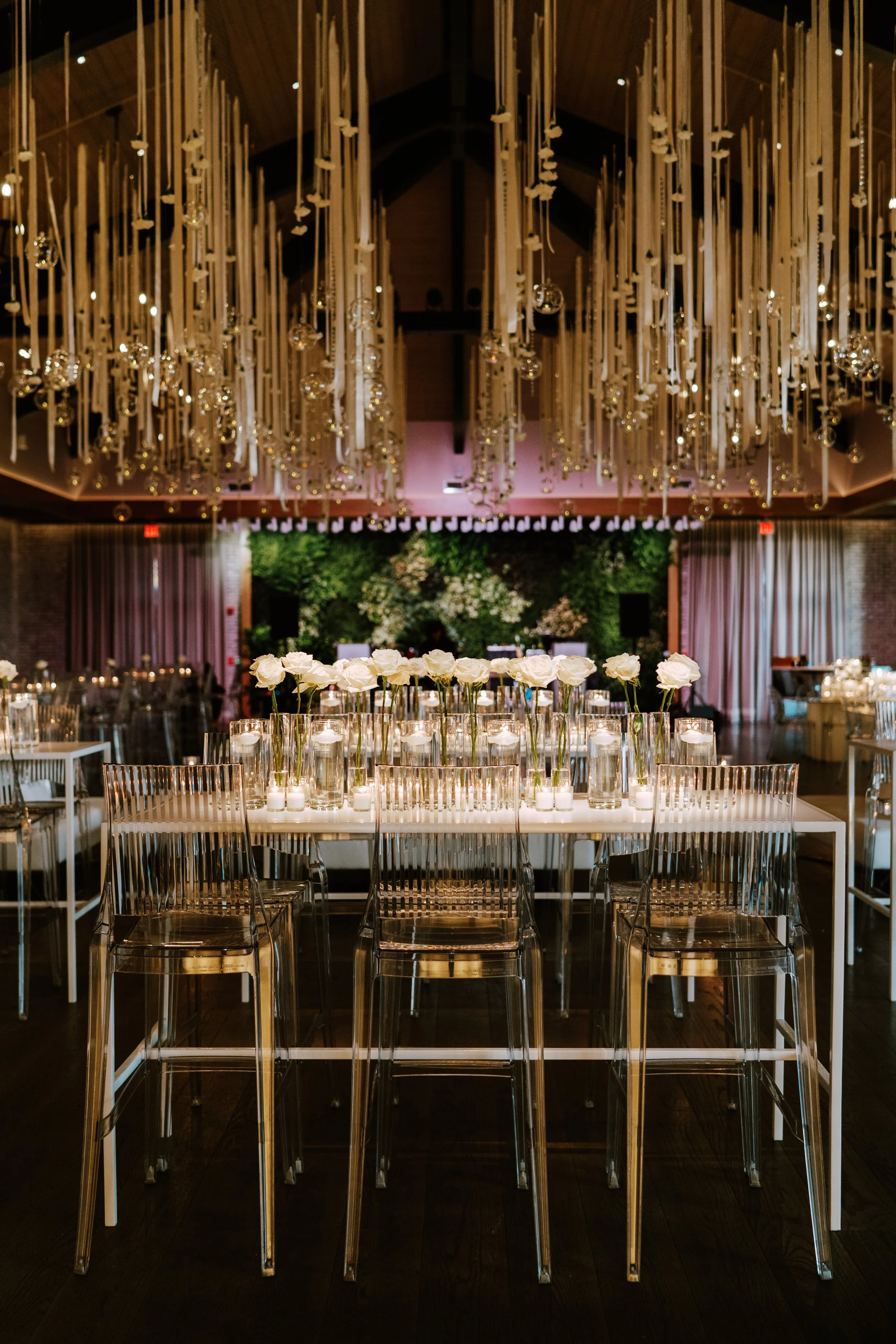 Elegant event setup with a long table decorated with white roses, candleholders, and candles, surrounded by transparent chairs, with hanging decorative elements and a green floral backdrop in a dimly lit venue.