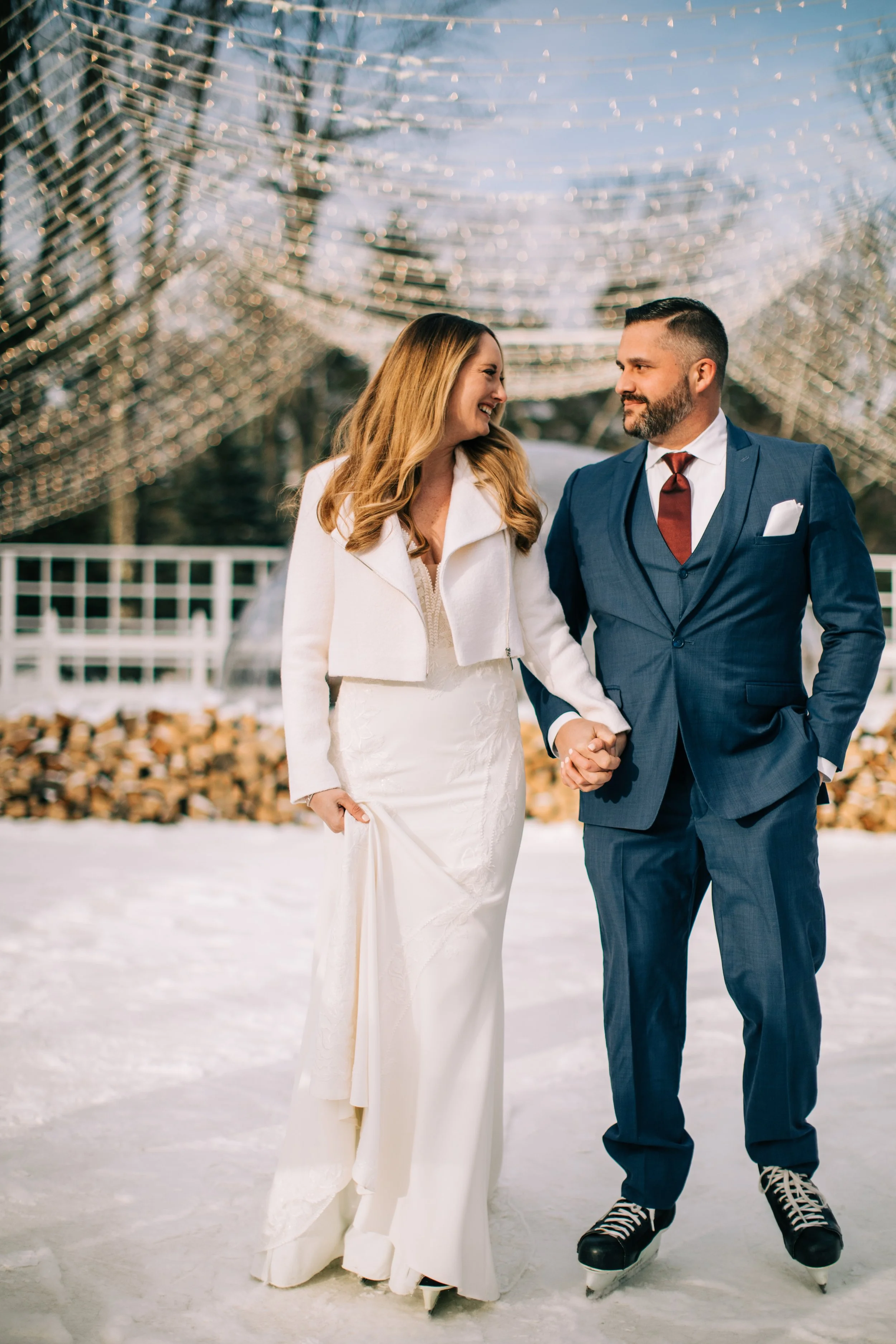 A bride and groom holding hands and smiling on an outdoor ice skating rink decorated with string lights, with a stack of firewood behind them.