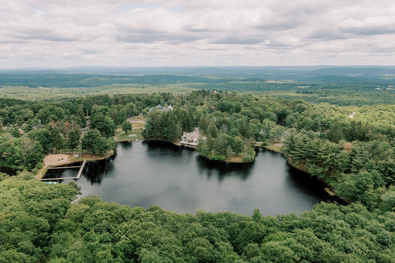 Aerial view of a large pond surrounded by dense green trees in a rural area with some houses and a small dock, under a cloudy sky.