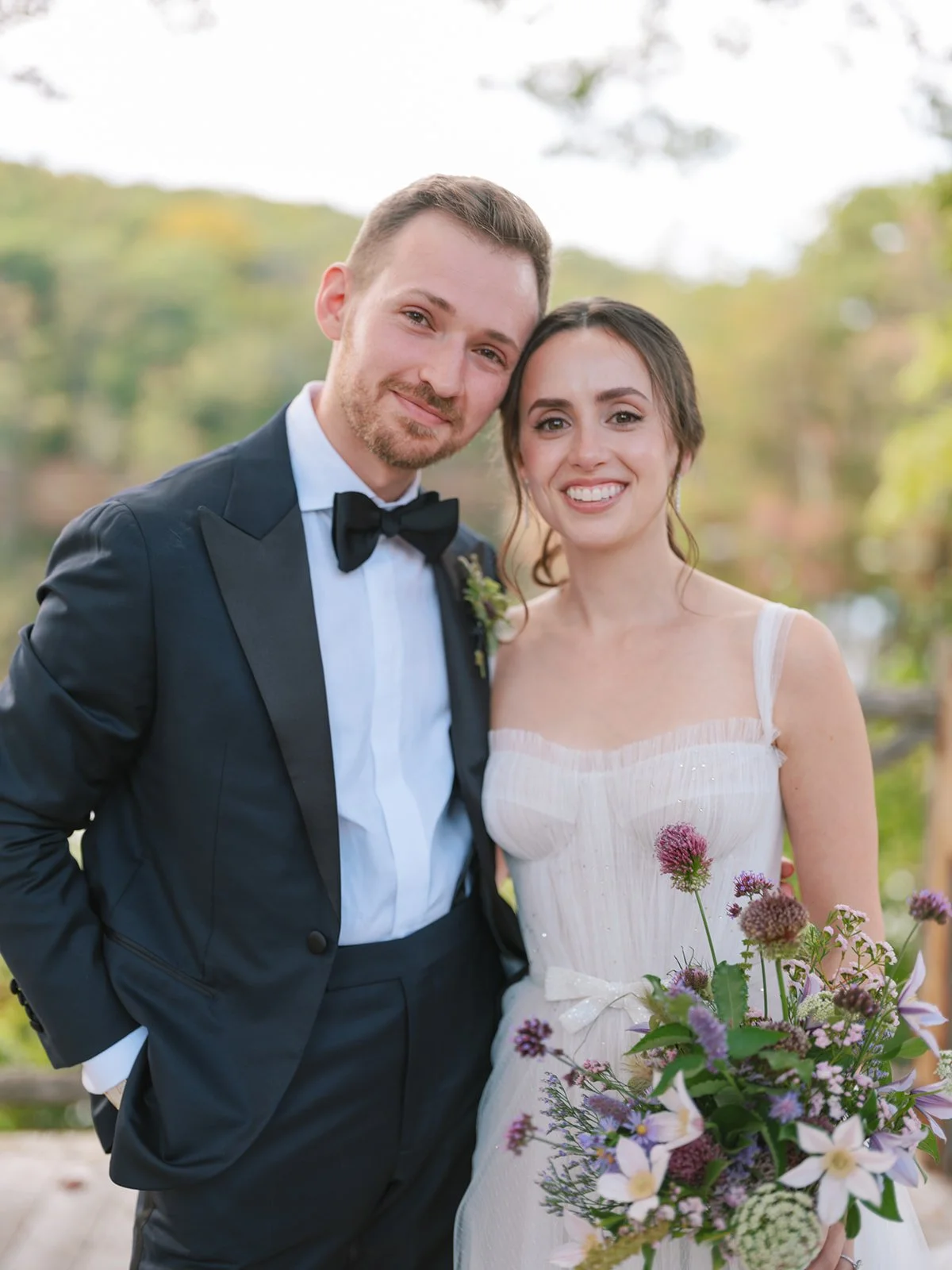 A newlywed couple dressed in formal wedding attire, standing outdoors with trees in the background, smiling at the camera. The groom wears a tuxedo with a black bow tie, and the bride wears a white dress holding a bouquet of colorful flowers.