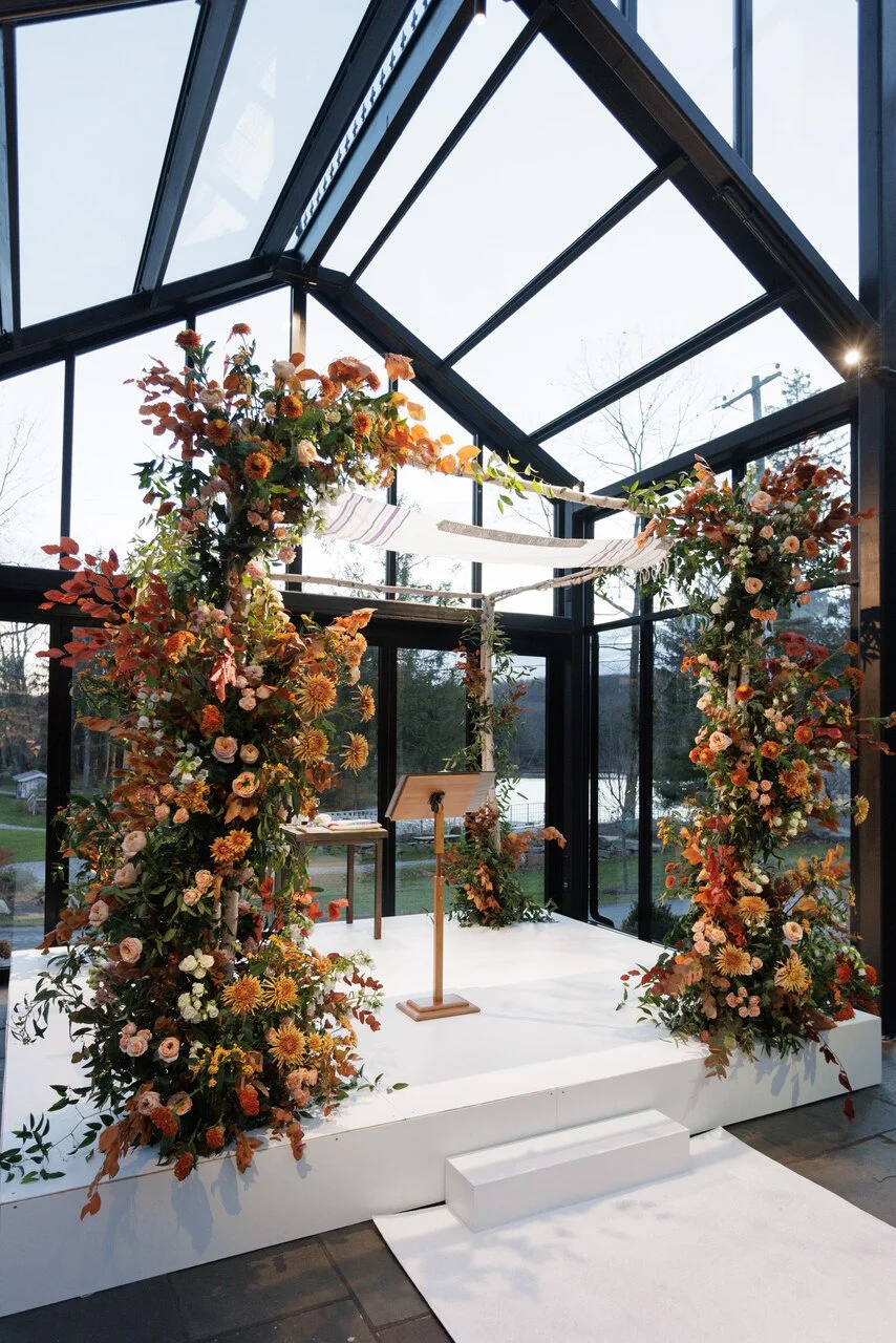 Floral wedding arch decorated with orange, peach, and pink flowers inside a glass conservatory