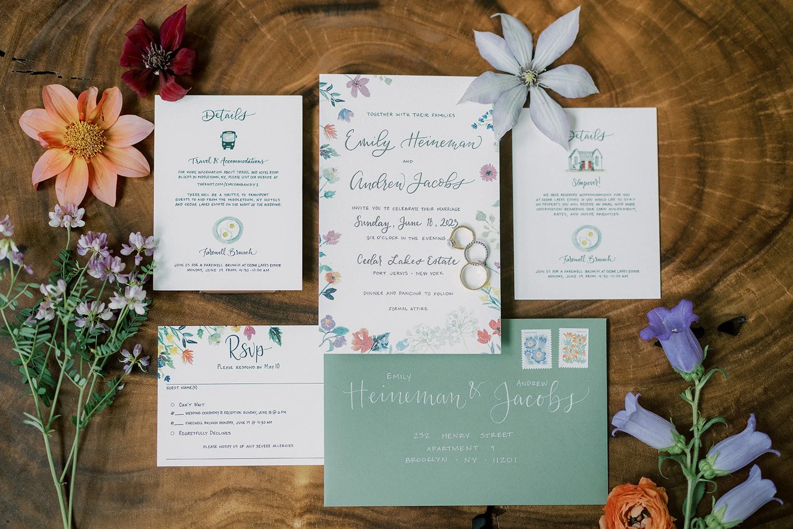 Wedding invitation suite surrounded by colorful flowers on a wooden surface. The set includes an invitation, RSVP card, details cards, and an envelope addressed to Emily Heineman & Andrew Jacobs.