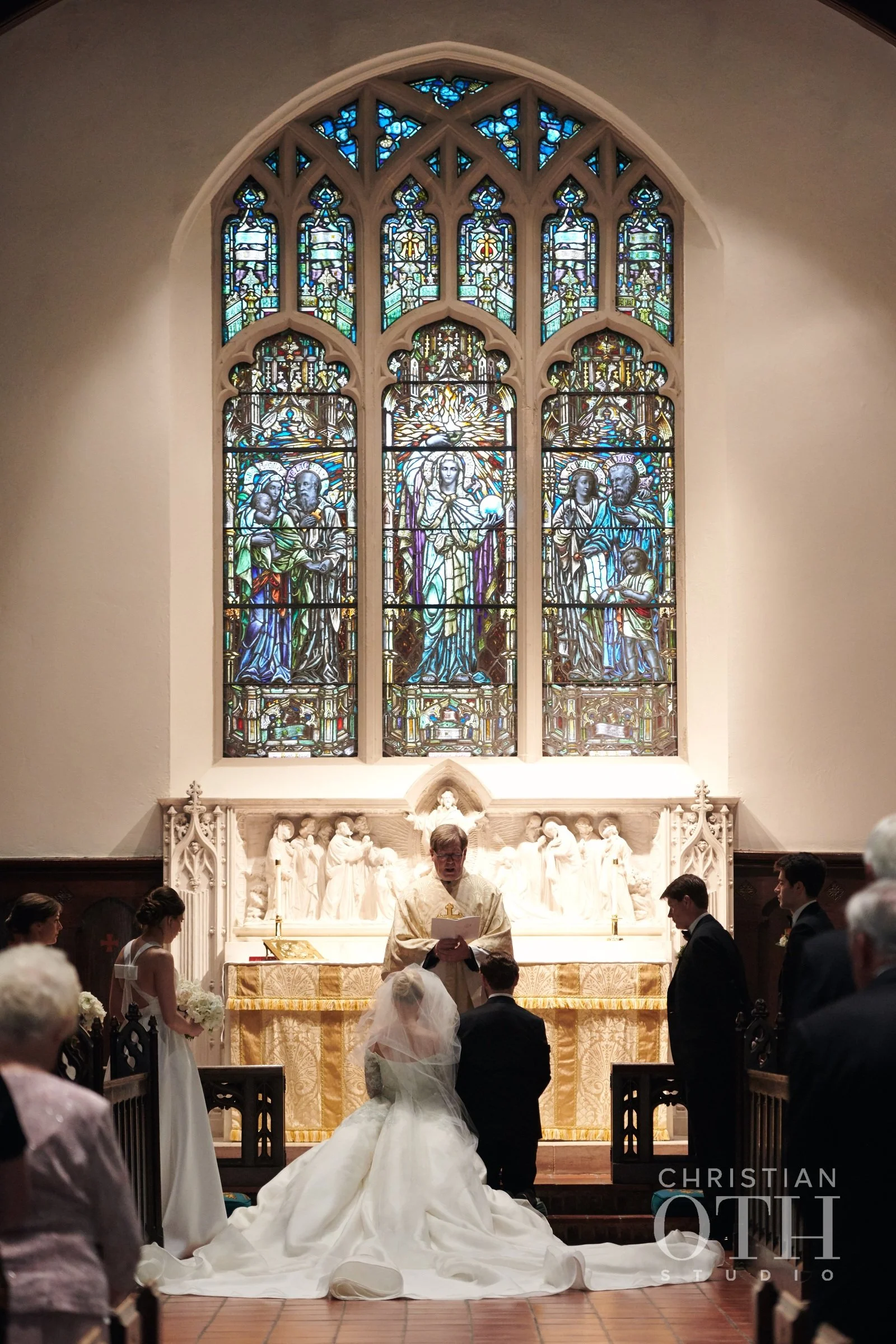 A wedding ceremony taking place in a church with a stained glass window depicting religious figures, and a priest officiating as the couple kneels before him, surrounded by guests.