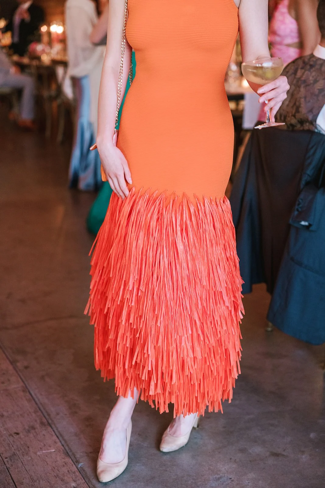 A woman in an orange dress with a fringed lower half, holding a glass of champagne at a social event.