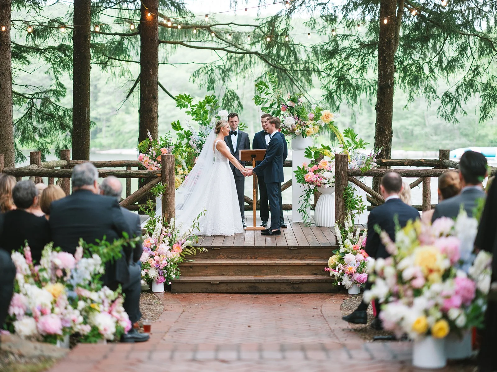 A wedding ceremony outdoors in a forested area with two people holding hands and exchanging vows. They are dressed in formal attire, with the bride in a white gown and the groom in a dark suit. An officiant stands behind them. The area is decorated w