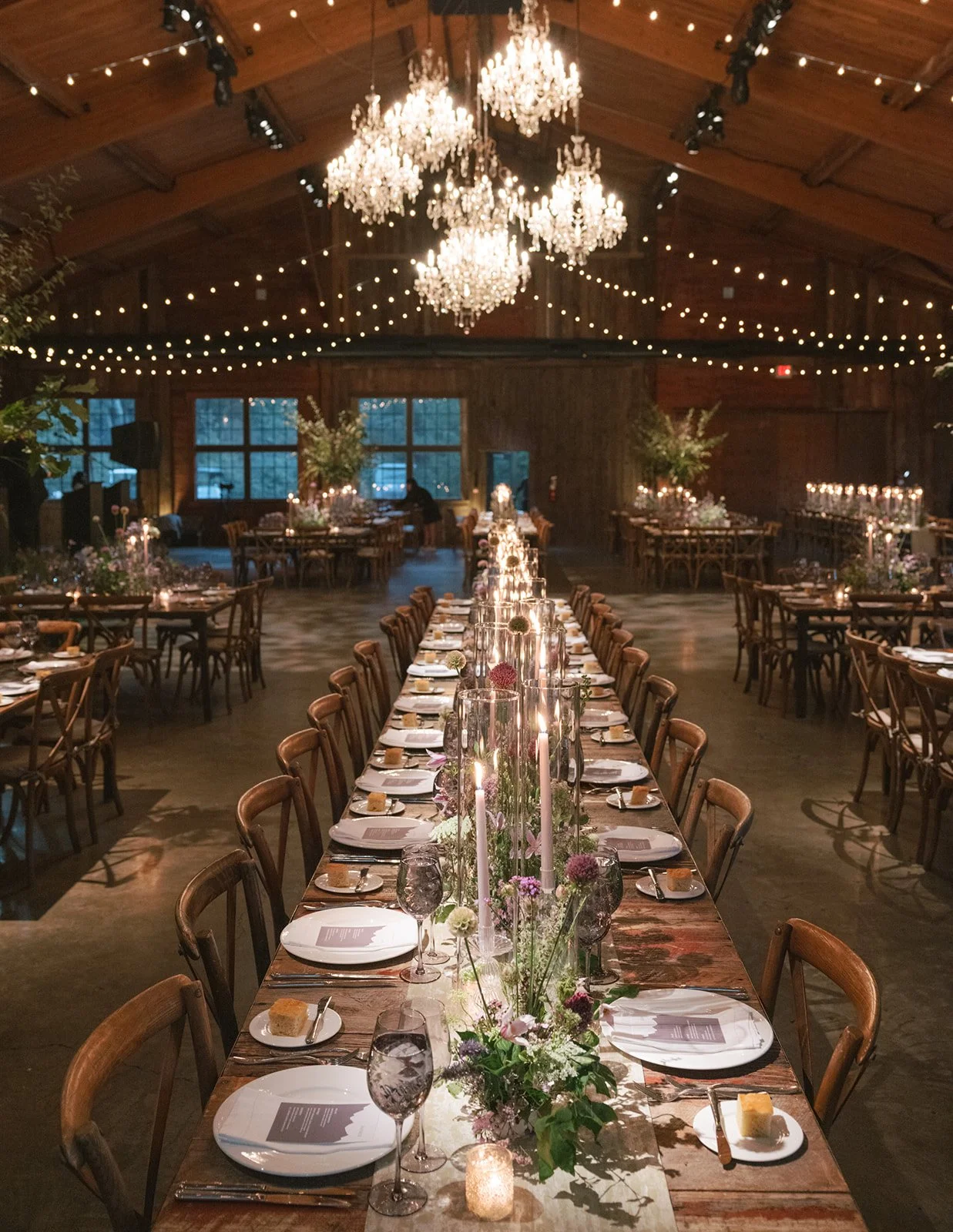 Wedding reception hall with long wooden table, floral centerpieces, tall candles, and hanging chandeliers and string lights in a rustic barn setting.