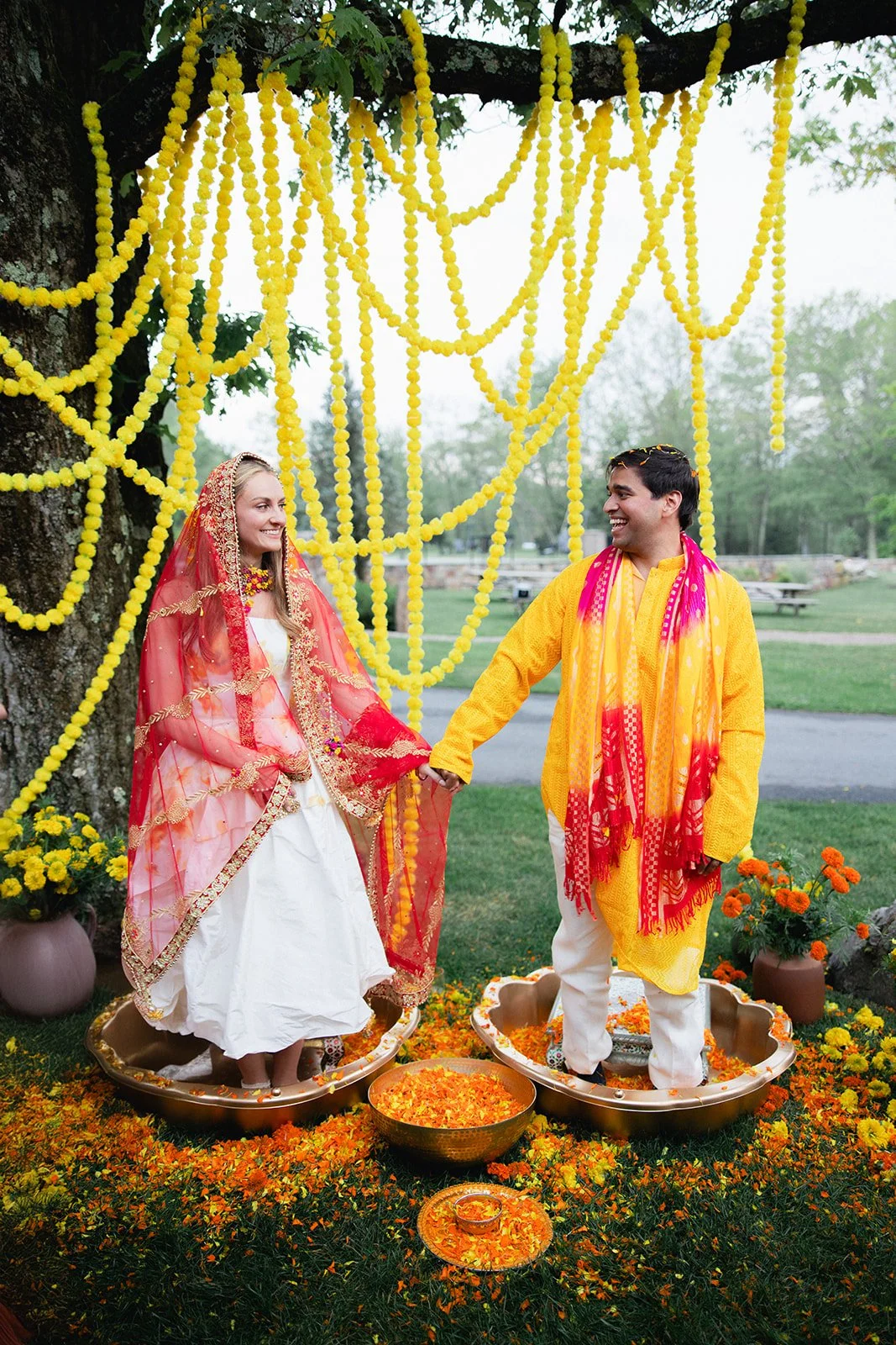 A couple dressed in traditional Indian attire participating in a cultural ceremony outdoors, standing on metal plates surrounded by flower petals, with yellow marigold decorations hanging from a tree behind them.