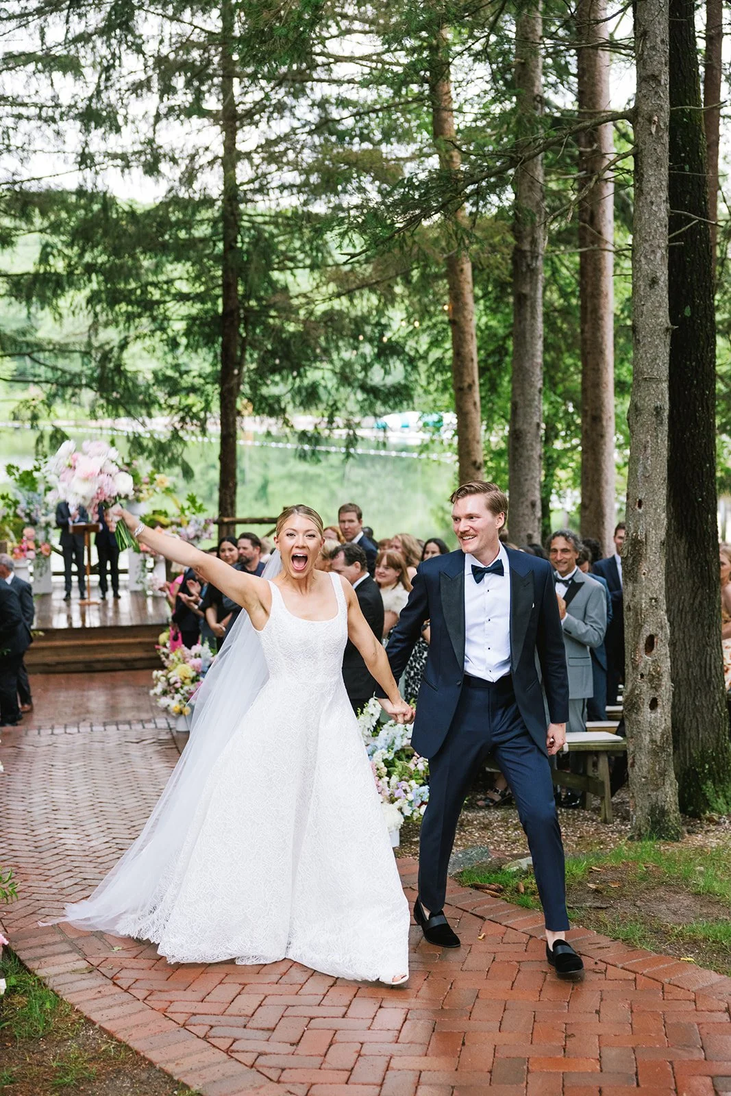A newlywed couple celebrates during their wedding ceremony outdoors in a wooded area, with guests standing and smiling in the background.