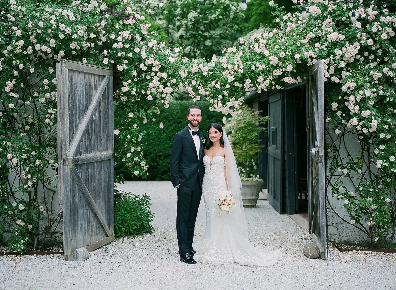 A bride and groom standing under an archway of blooming pink and white roses on their wedding day, smiling at the camera.