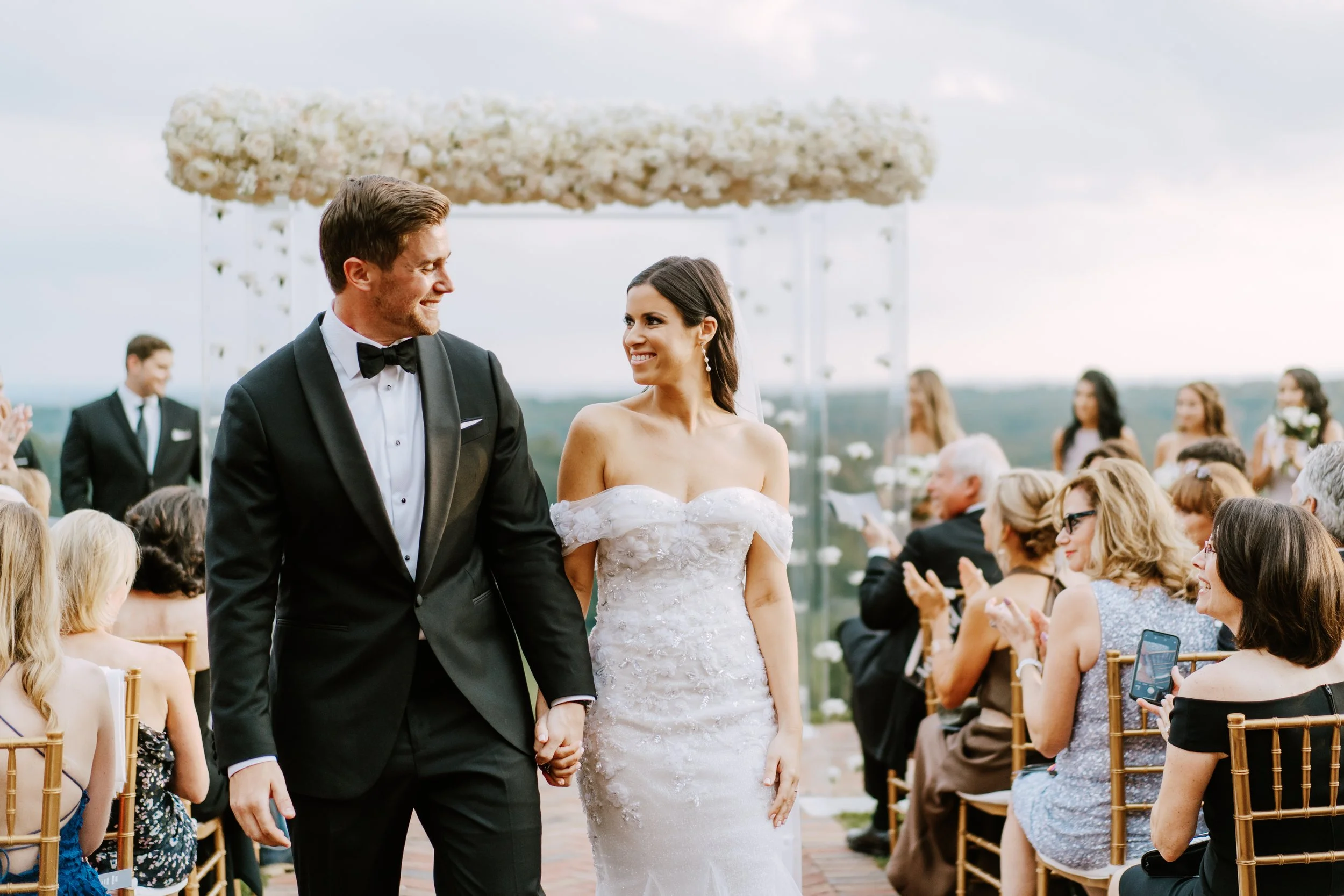 A bride and groom holding hands, smiling, during their outdoor wedding ceremony with guests seated around them.