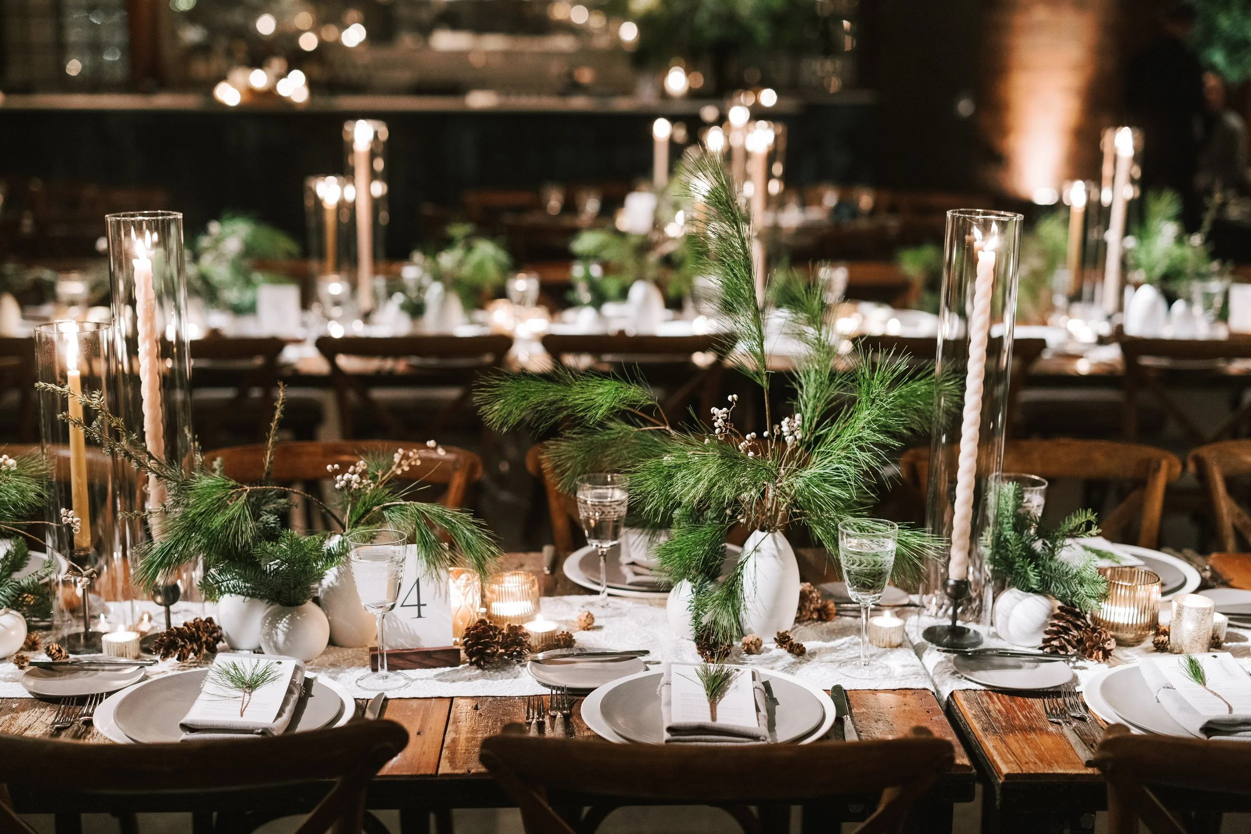 Festively decorated banquet table with green pine branches, candles, pinecones, and white tableware, set for a holiday celebration.