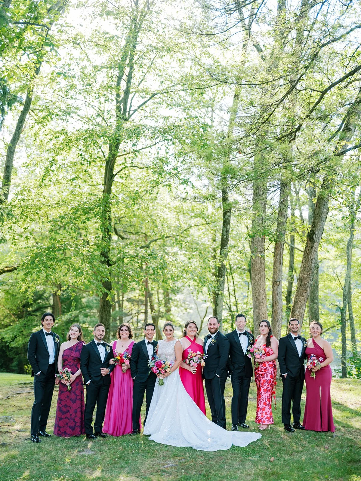A wedding party standing outdoors on grass, with trees in the background. The group includes a bride in a white wedding dress, bridesmaids in pink dresses, and groomsmen in black tuxedos. They are all smiling and holding bouquets.