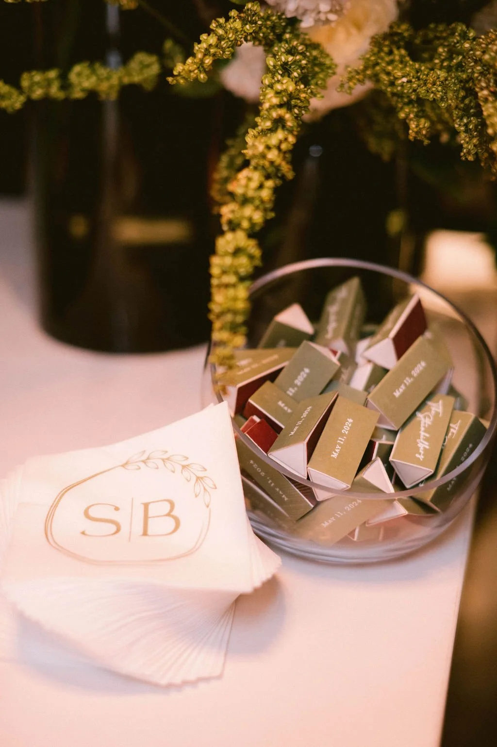 A stack of napkins with a monogram 'SB' in gold, and a glass bowl filled with small rectangular favor boxes with dates and wedding initials printed on them, placed on a table with floral decorations in the background.