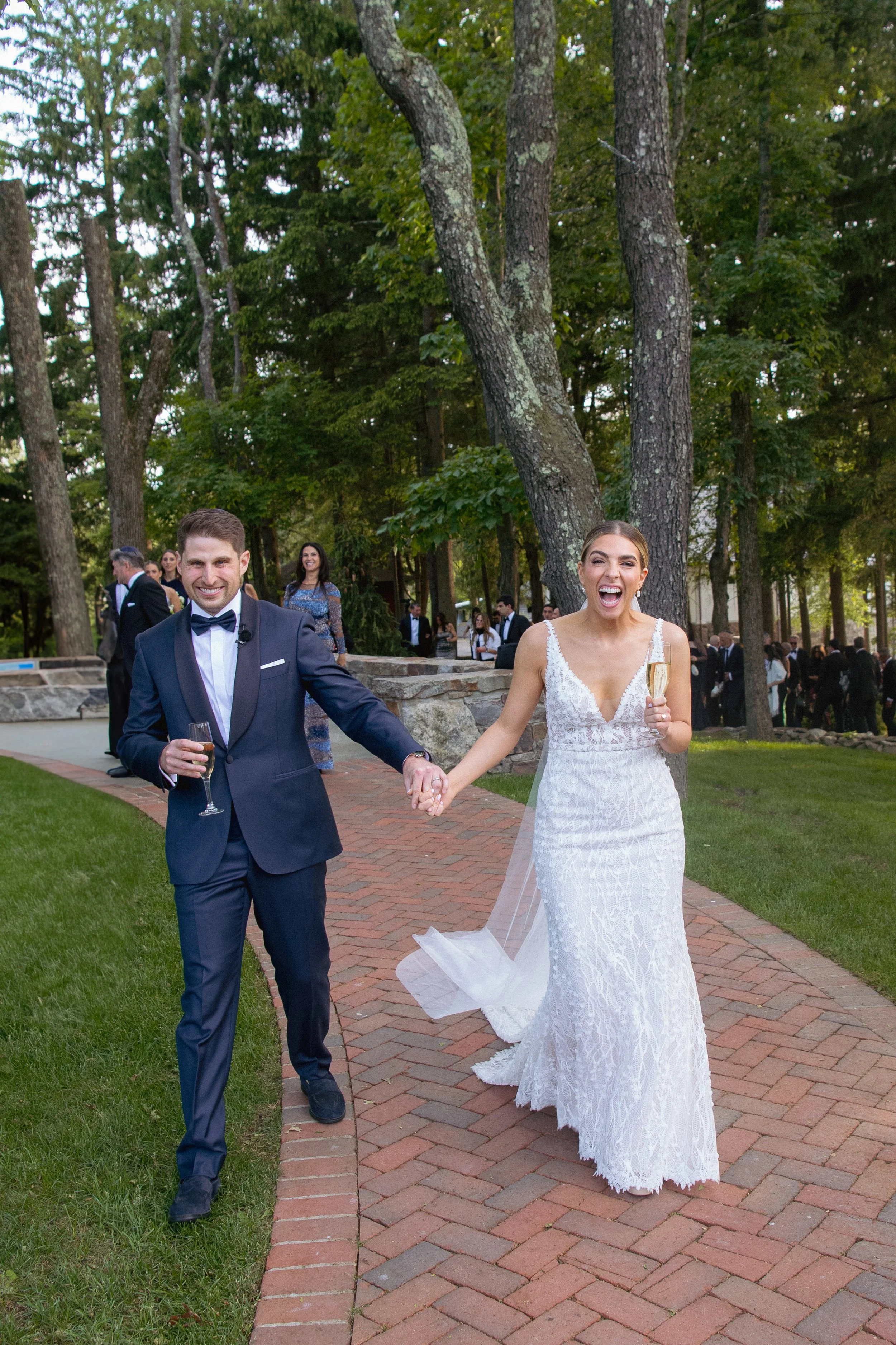 A wedding celebration outdoors with a bride and groom walking hand in hand, both holding champagne glasses, smiling, and surrounded by guests in formal attire in a wooded area.