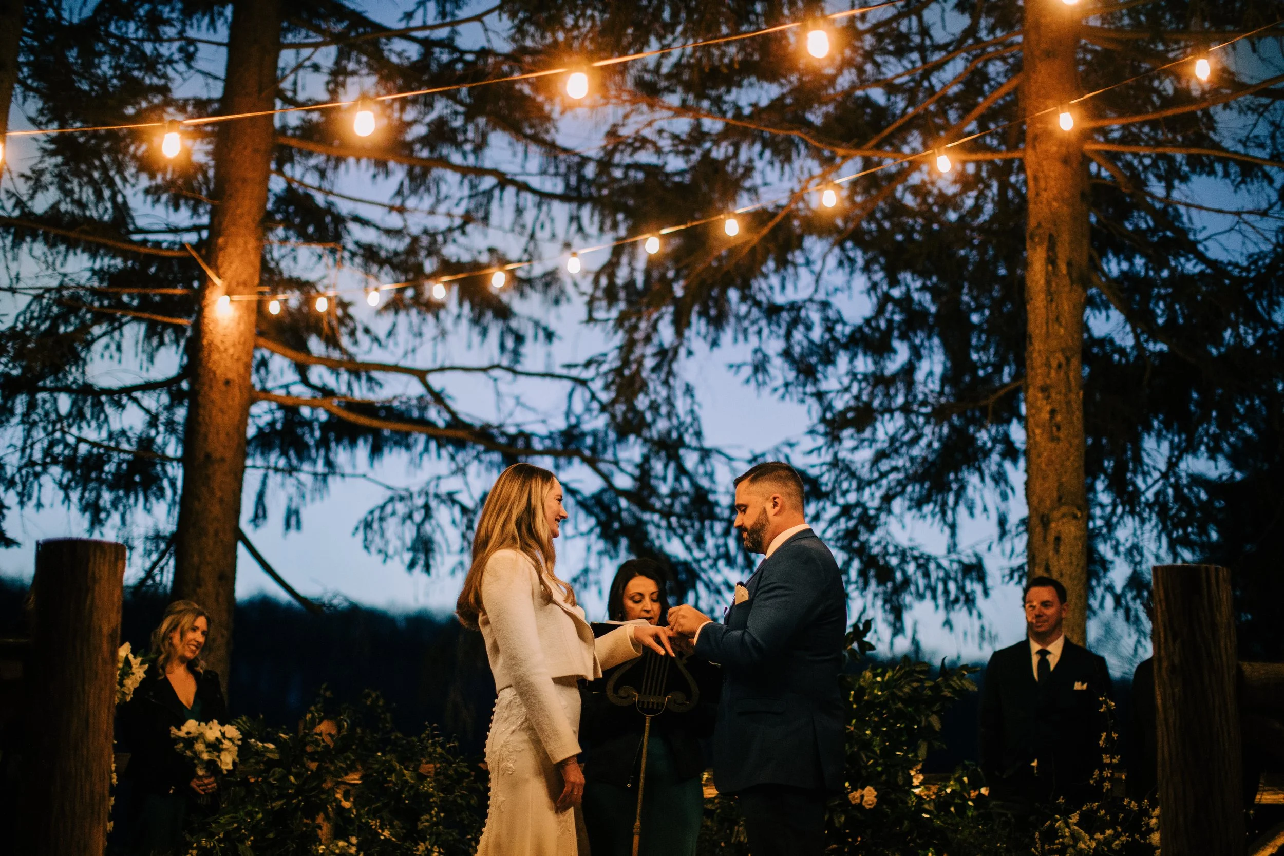 A wedding ceremony outdoors at night under string lights, with a bride and groom exchanging rings, surrounded by bridesmaids and groomsmen, with trees and decorations in the background.