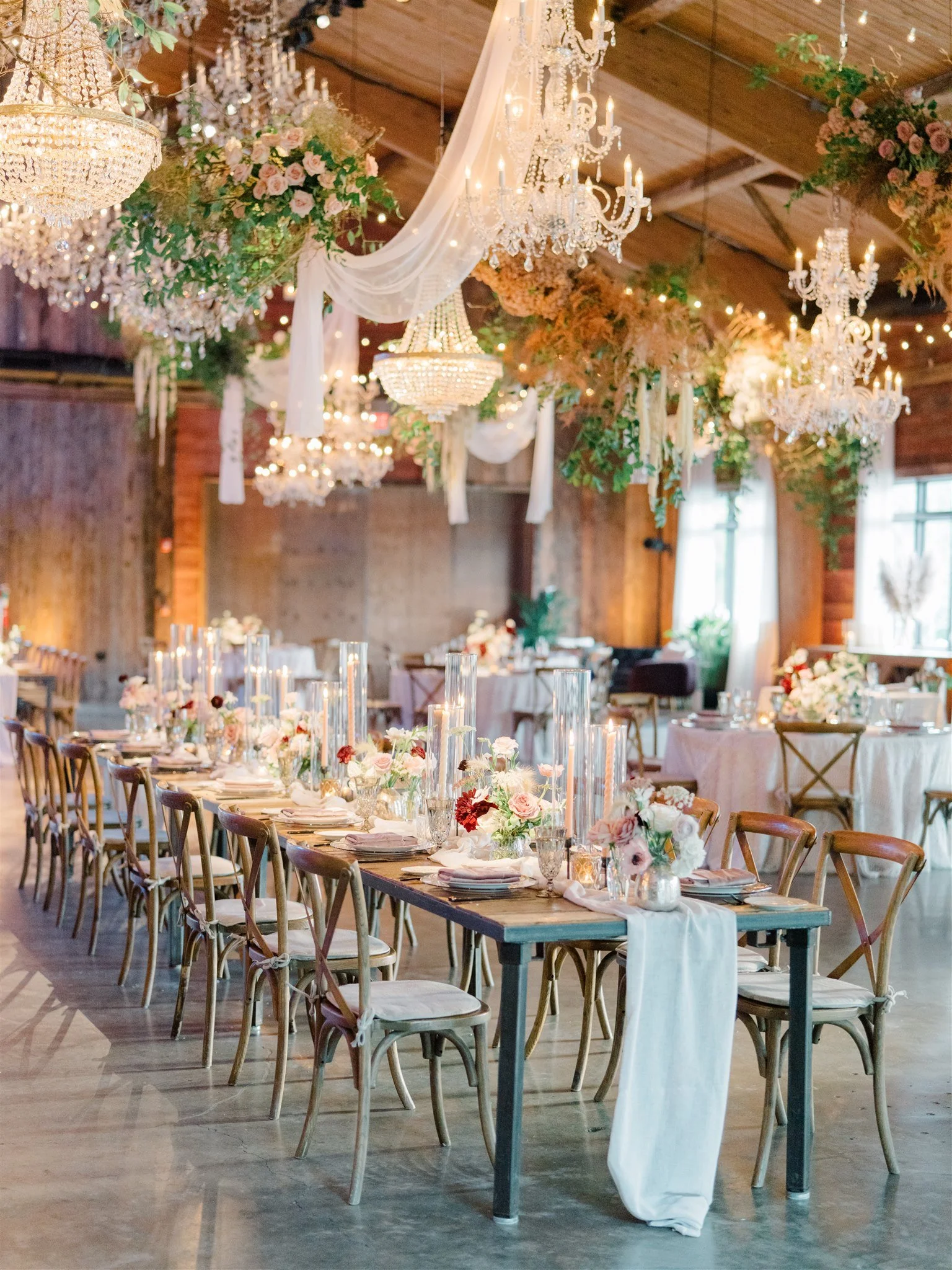 Elegant wedding reception with long wooden table decorated with pink and white flowers and tall glass candle holders, chandeliers, floral arrangements, and soft lighting in a rustic venue with wooden walls and ceiling.