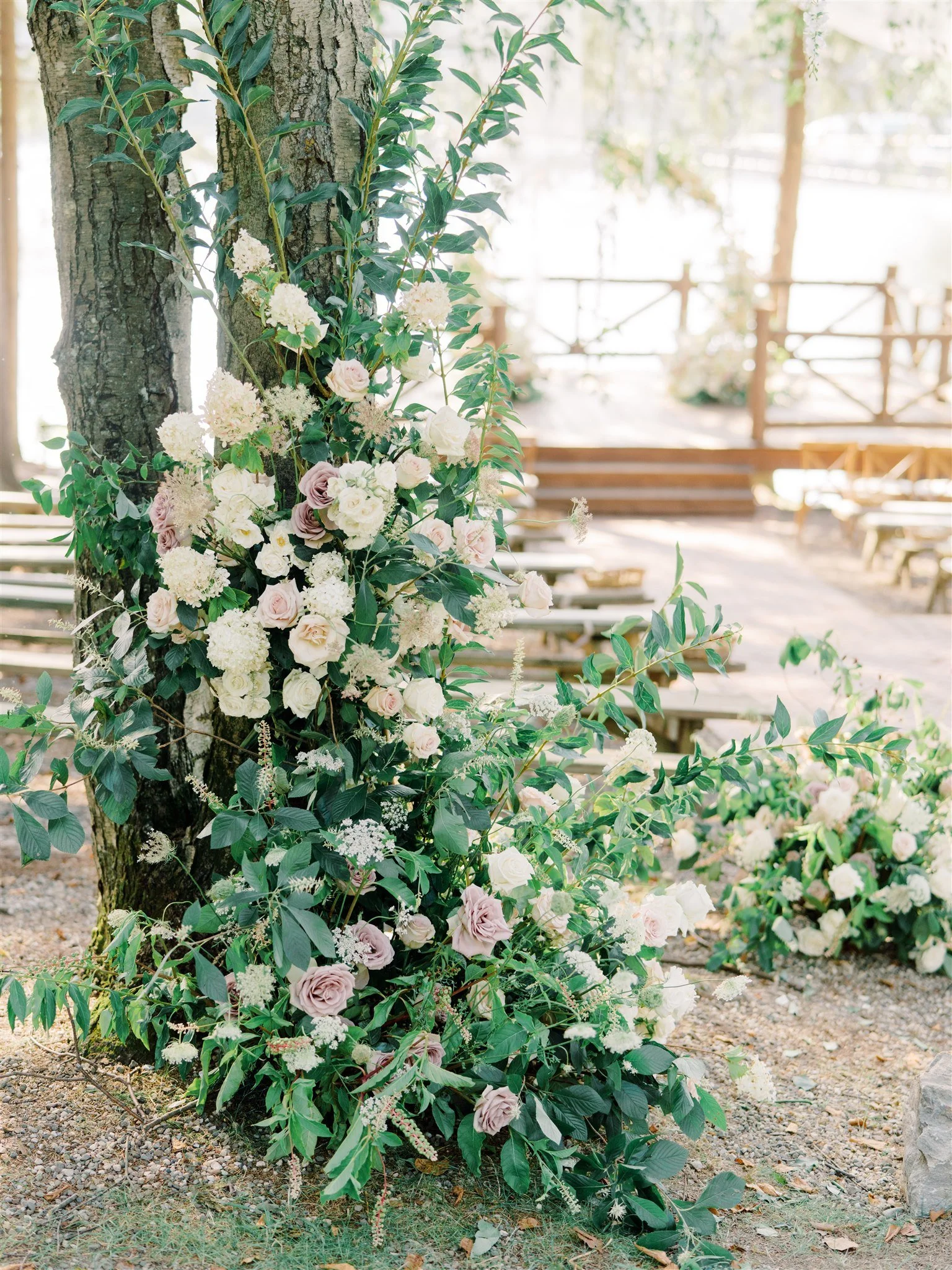Floral decoration with white and soft pink roses, hydrangeas, and lush green foliage arranged around a tree trunk outdoors, likely for a wedding or special event.