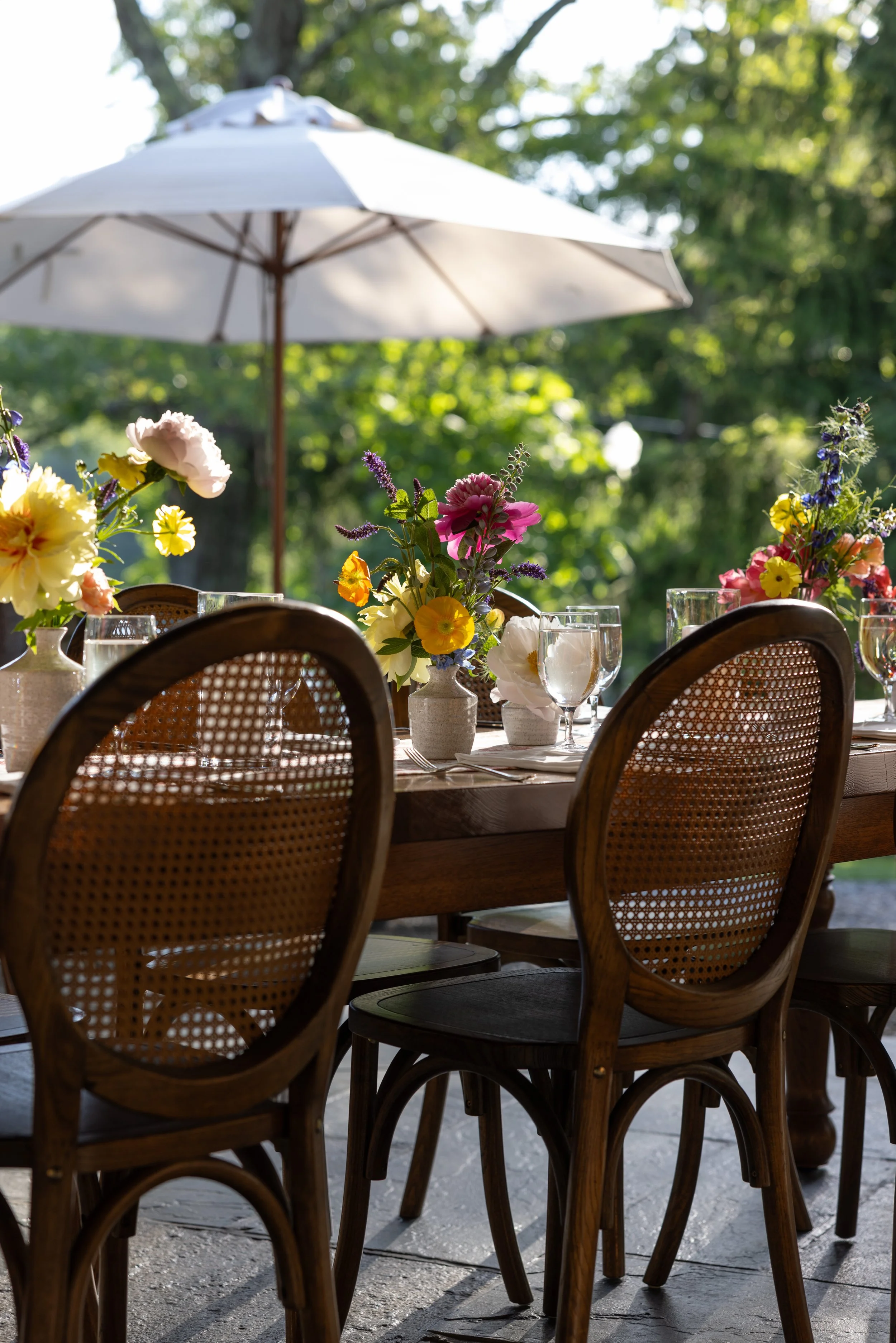An outdoor dining table set with colorful flower centerpieces, glassware, and cutlery, shaded by a large white patio umbrella with green trees in the background.