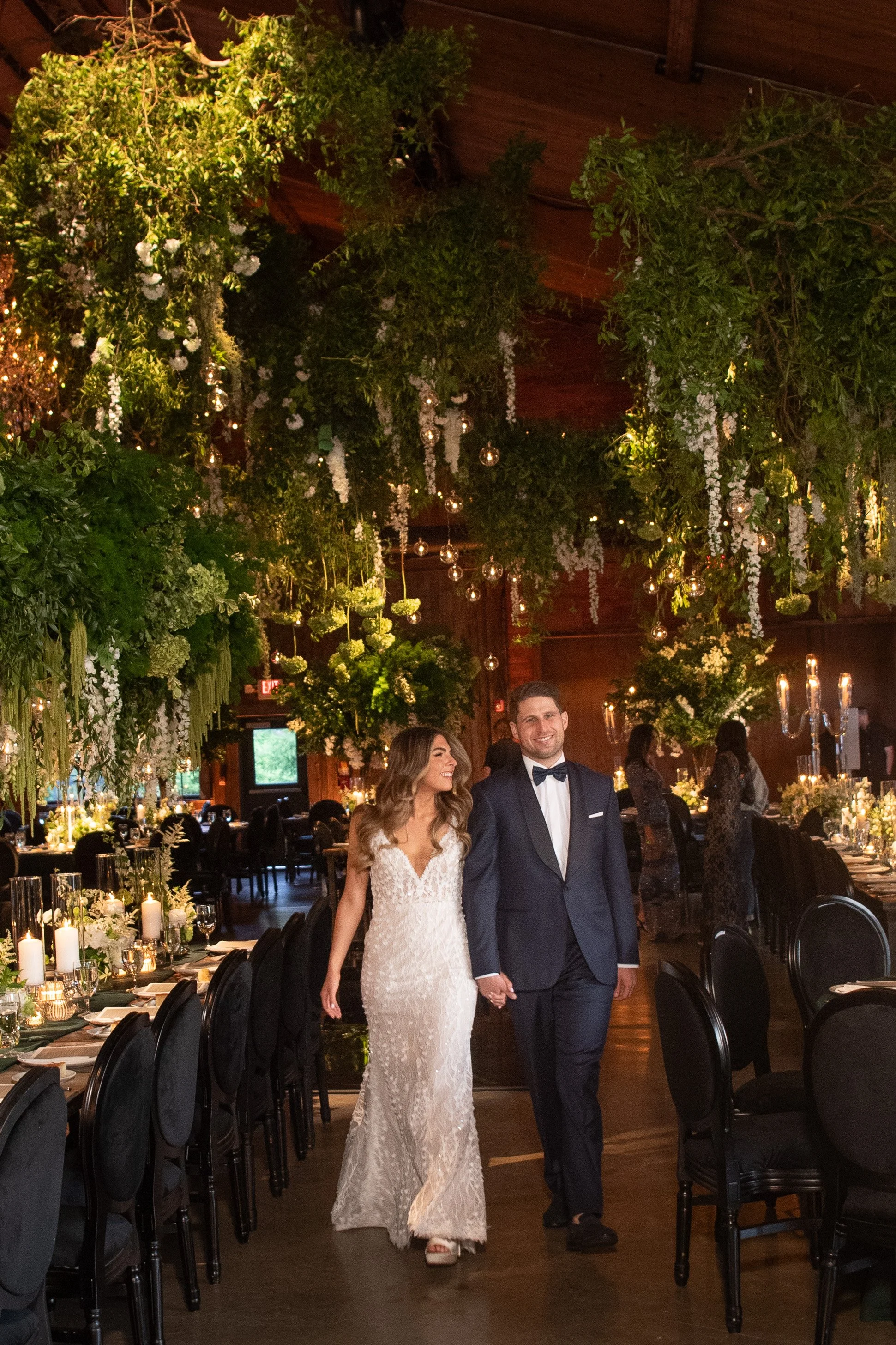 A couple dressed in wedding attire, holding hands and walking through a decorated wedding reception hall with hanging greenery, white flowers, candles, and formal table settings.