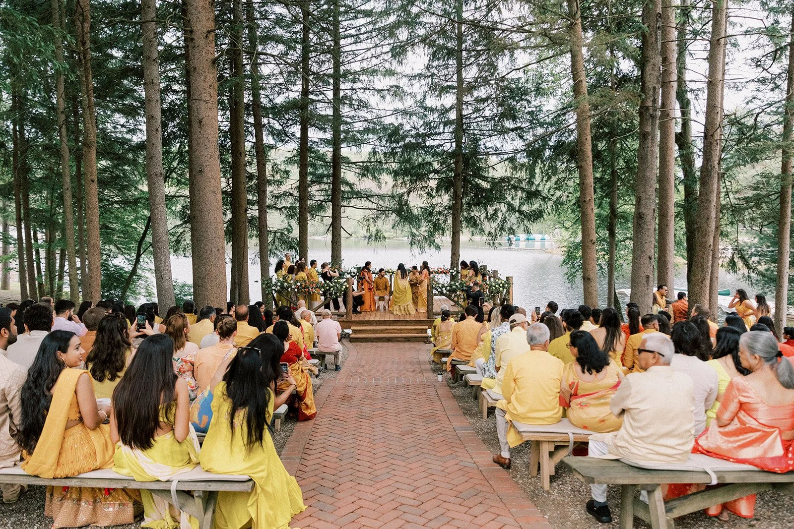 A large outdoor gathering by a lake features a wedding ceremony with guests seated on benches in front of a decorated altar, surrounded by tall pine trees.