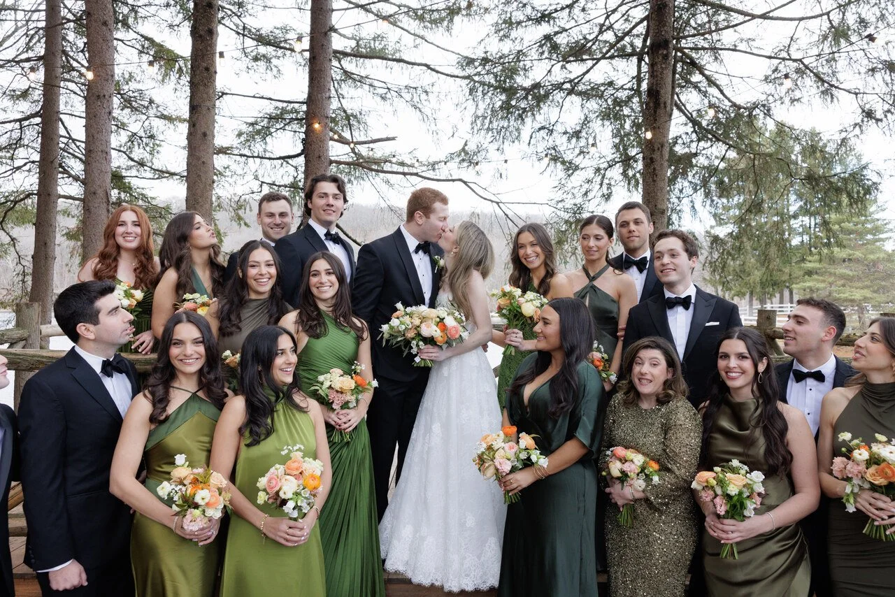 Group of wedding guests and the bride and groom outdoors with trees in the background, some holding bouquets, celebrating at a wedding.