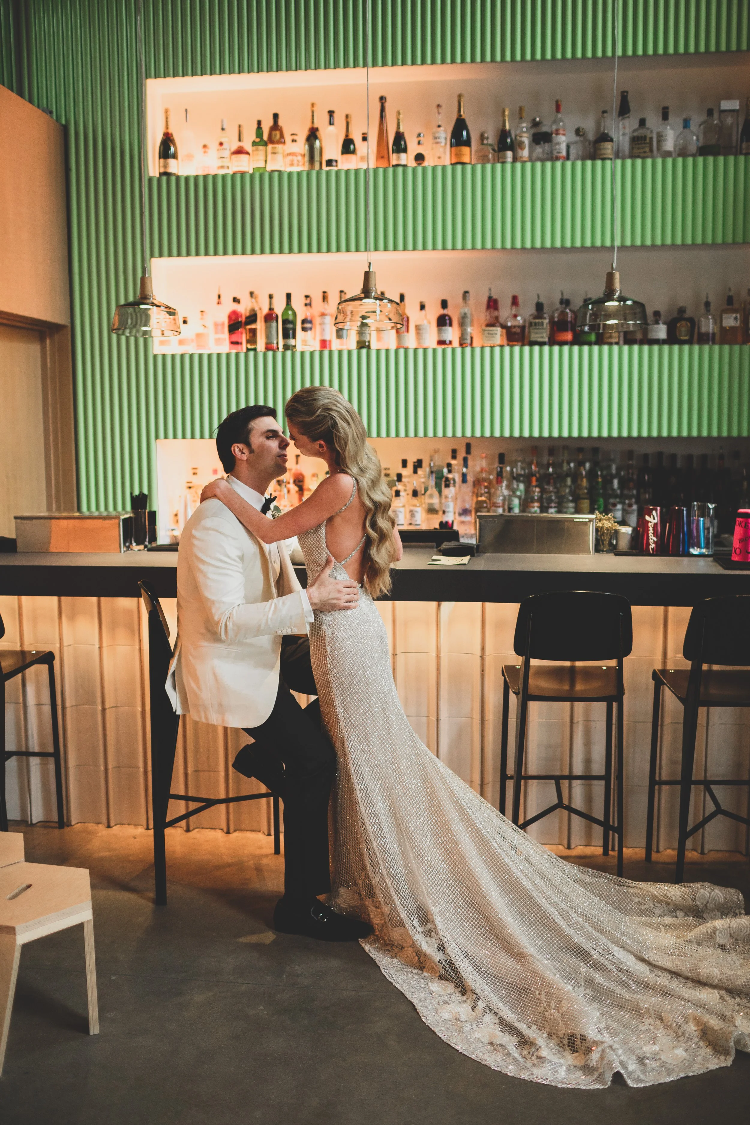 A newlywed couple shares a romantic moment at a bar. The groom is sitting on a barstool, wearing a white tuxedo jacket and black pants. The bride leans in close, wearing a sparkling, backless wedding gown with a long train. The bar has a colorful, mo