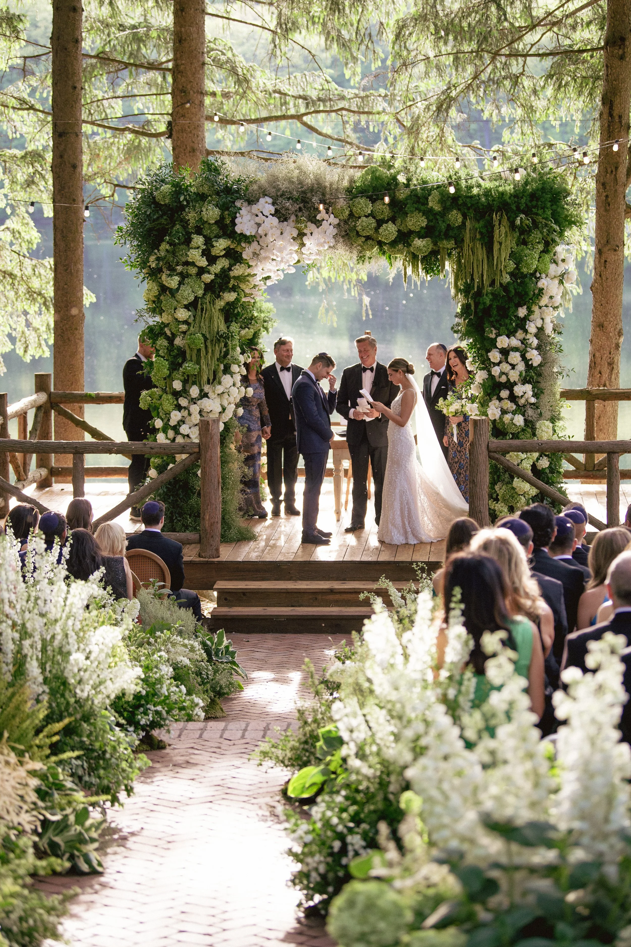 A wedding ceremony taking place under a floral arch on an outdoor wooden platform, with the bride and groom standing in front of an officiant, surrounded by seated guests and tall trees in the background.
