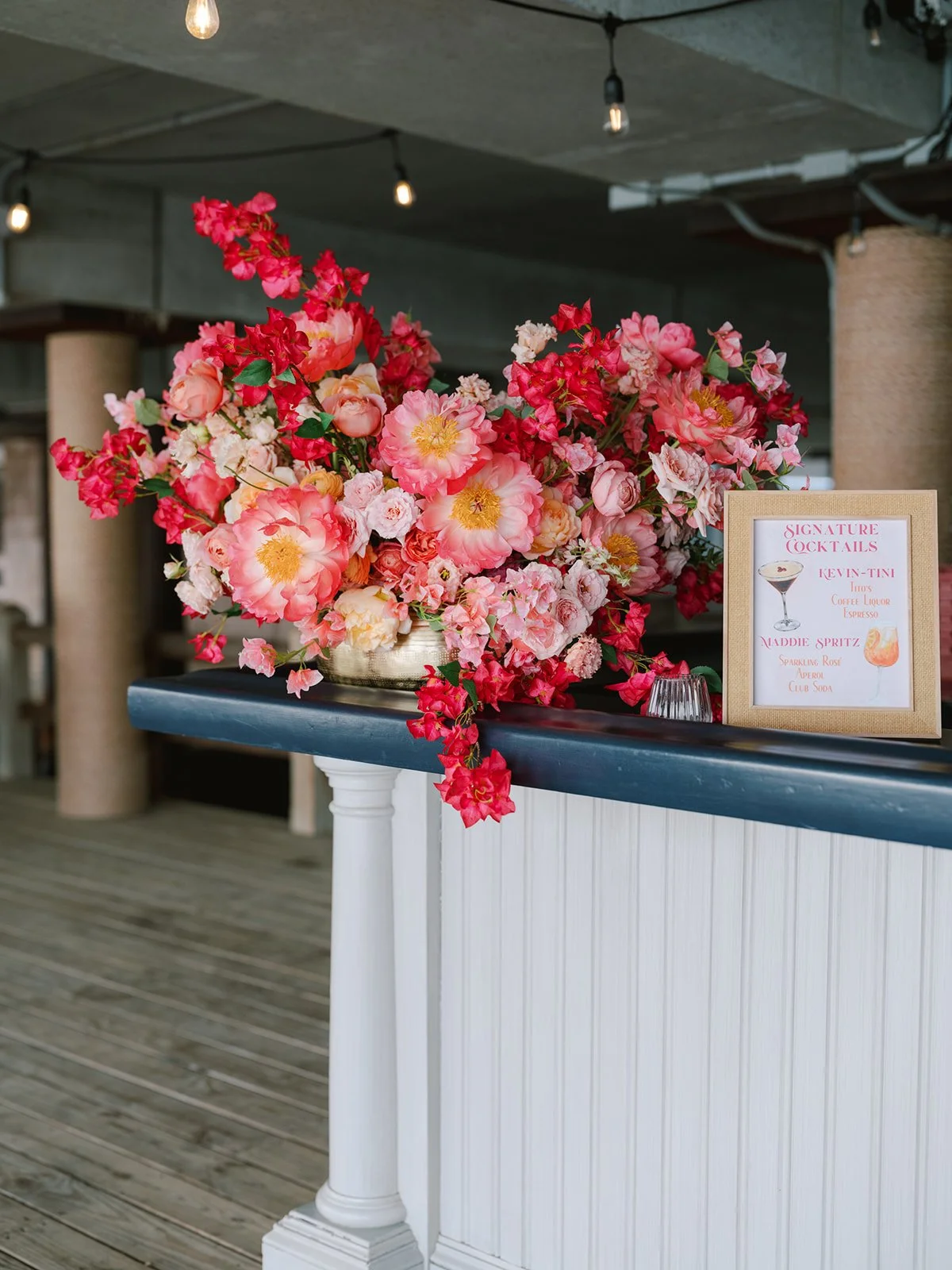 A colorful floral arrangement with pink, red, and white flowers on a dark table or countertop, with a sign listing signature cocktails nearby, in an indoor setting with string lights hanging from the ceiling.