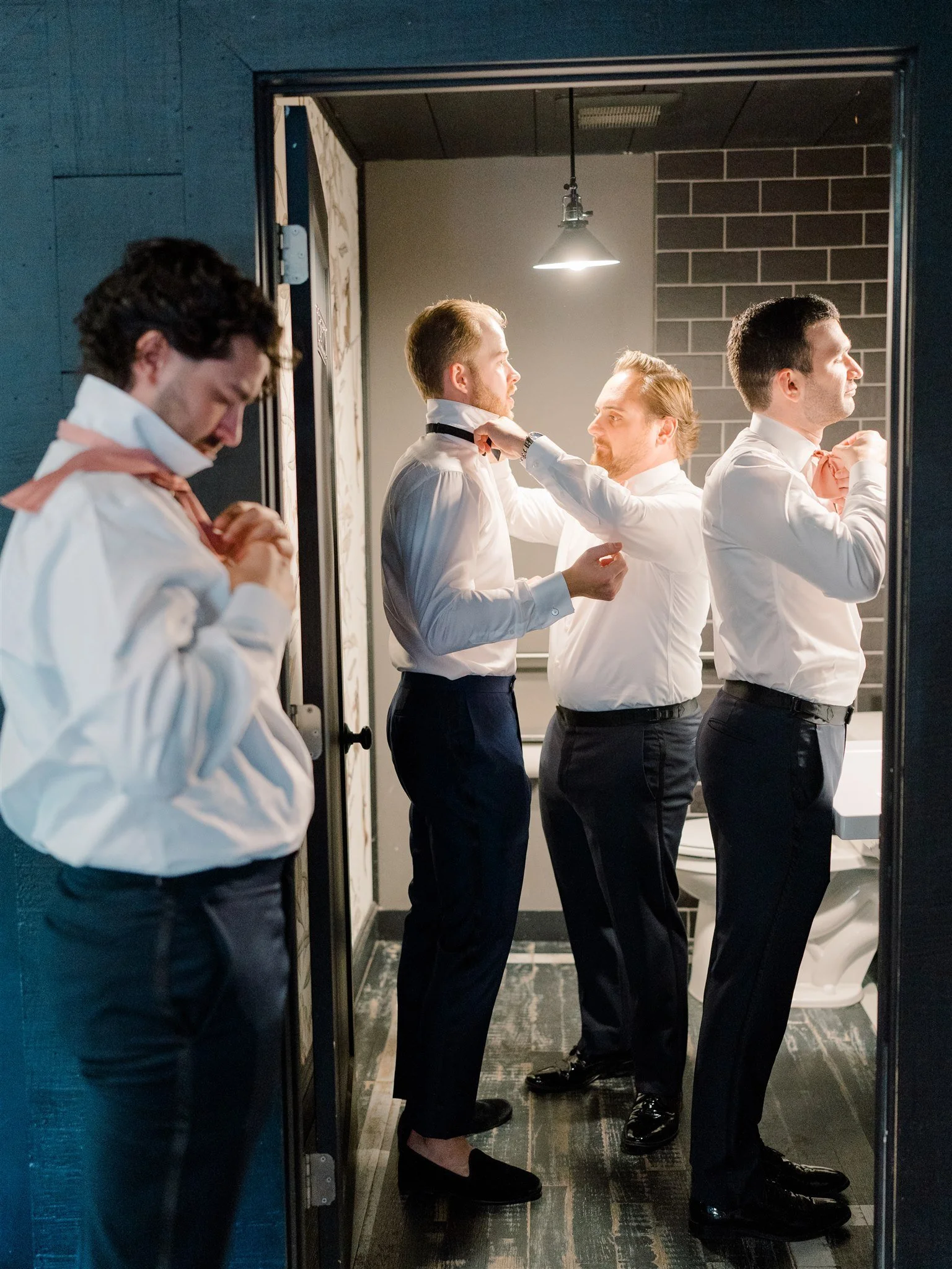 Four men getting dressed and ready in a bathroom, with three of them adjusting their ties and one doing something near his face.