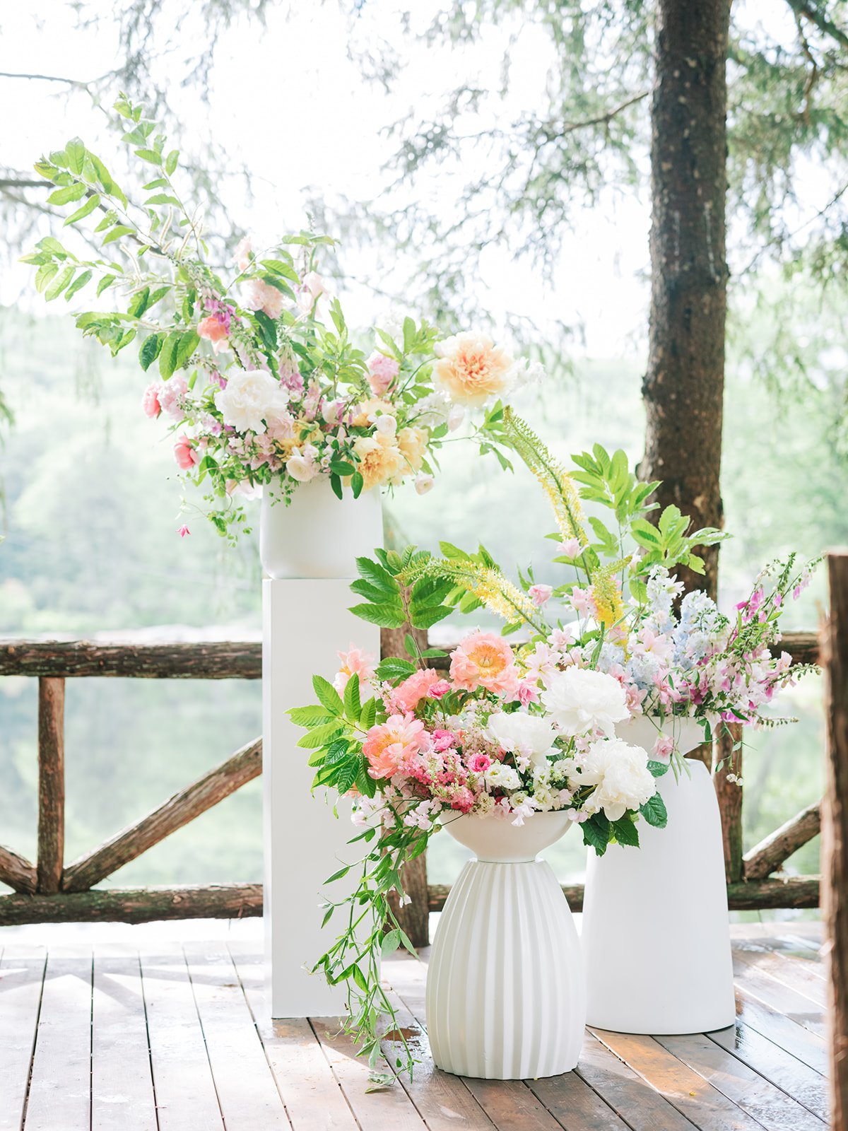 Vases with colorful flowers and greenery on a wooden deck with trees in the background.