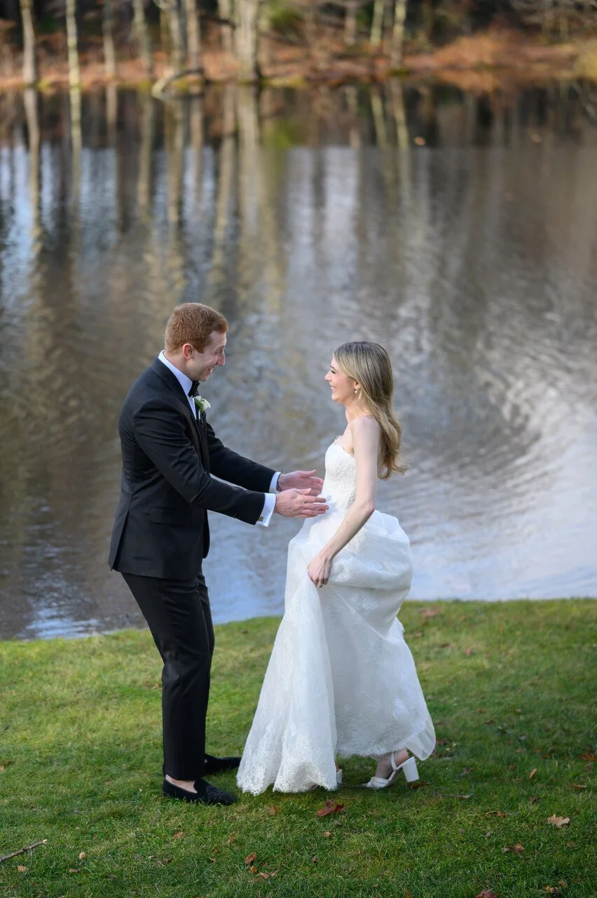 A bride and groom standing on grass by a pond, smiling and holding hands during their wedding.