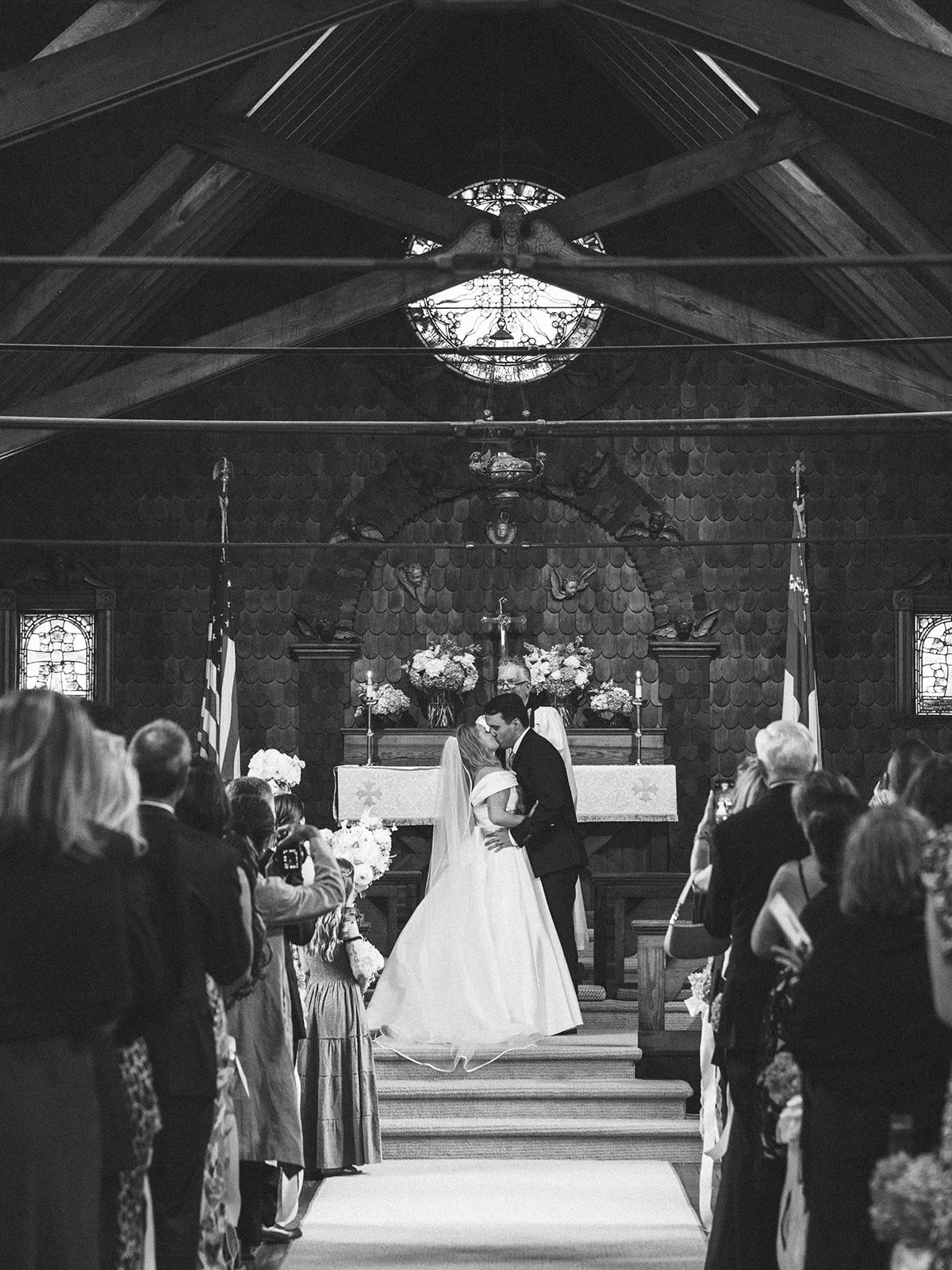A wedding ceremony inside a church with a couple kissing at the altar, surrounded by guests taking photos.