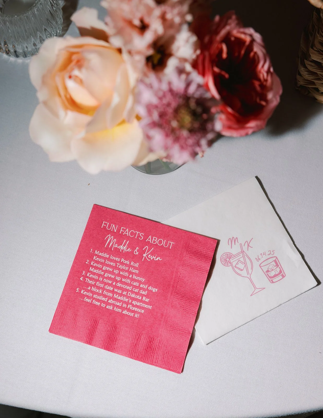 A bouquet of light pink, peach, and dark red flowers on a table next to a white napkin with a hand-drawn drink and date, along with a pink card titled 'Fun Facts About Maddie & Kevin' listing facts, and a white card with pink illustration.