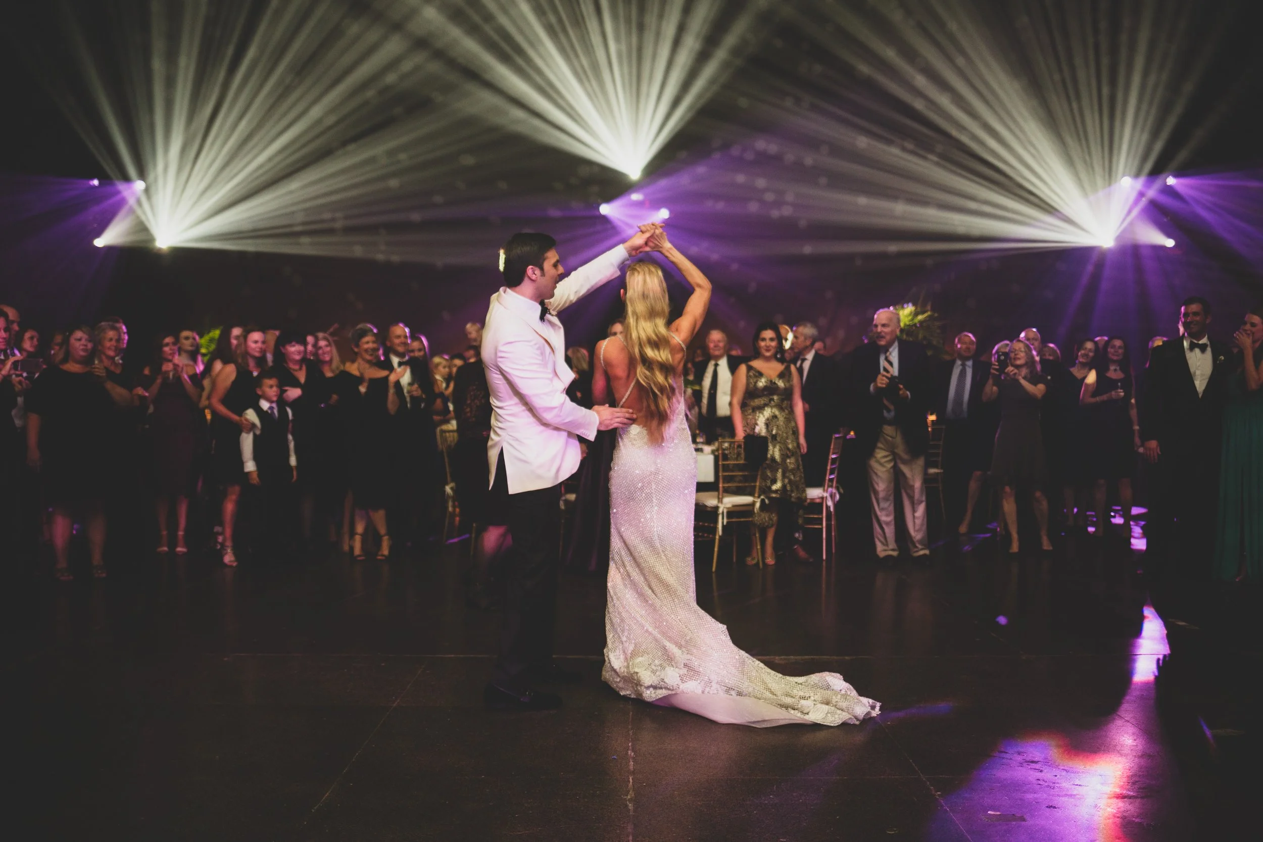 A bride and groom are dancing together during their wedding reception with guests watching and taking photos in the background, while colorful lights illuminate the dark dance floor.
