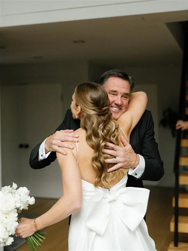 A bride and an older man, likely her father, sharing a warm embrace at her wedding. The bride has long, wavy hair and is holding a bouquet of white flowers.