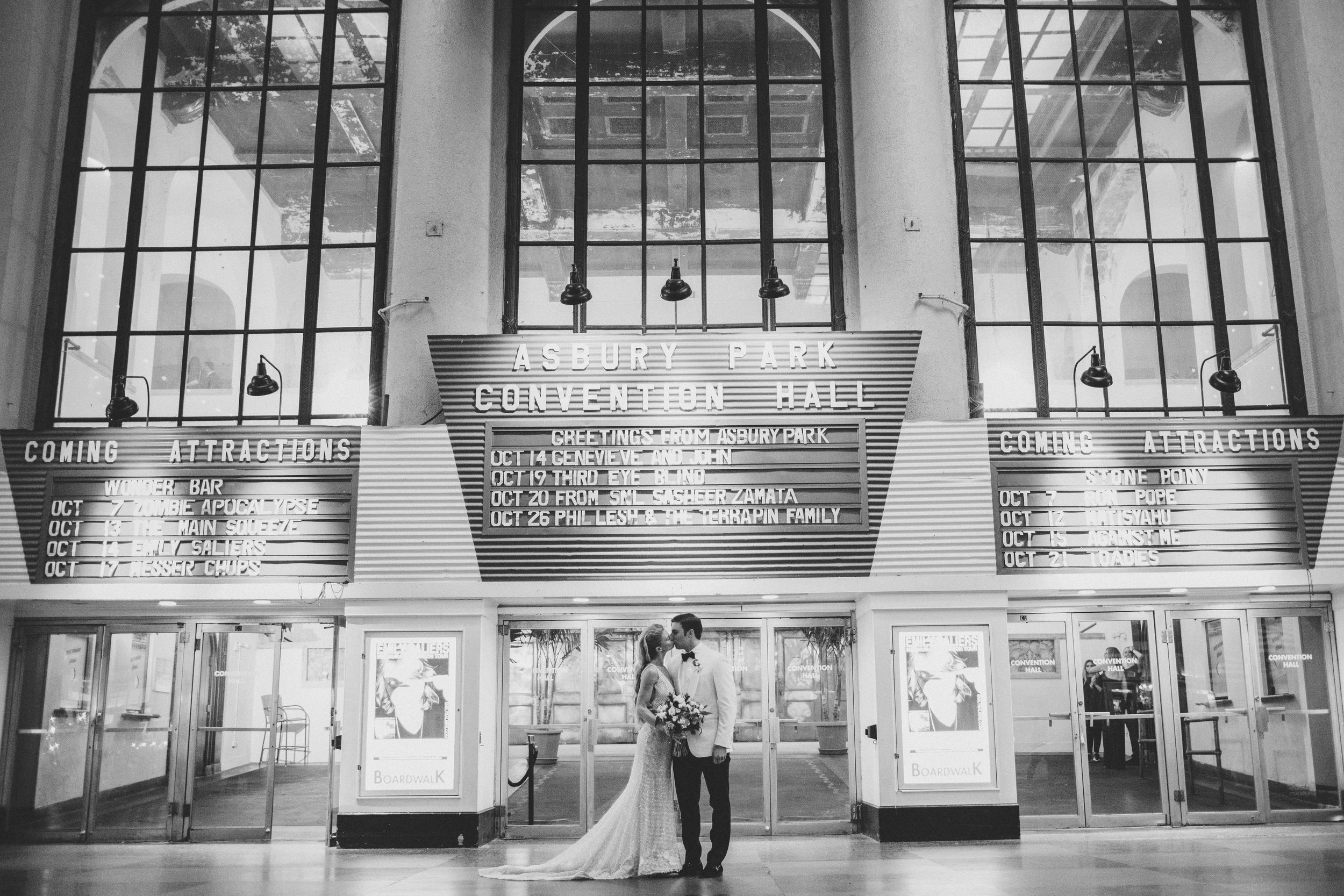 A bride and groom stand together in front of the entrance to Asbury Park Convention Hall, holding hands and smiling at each other, with large windows and brackets above them.