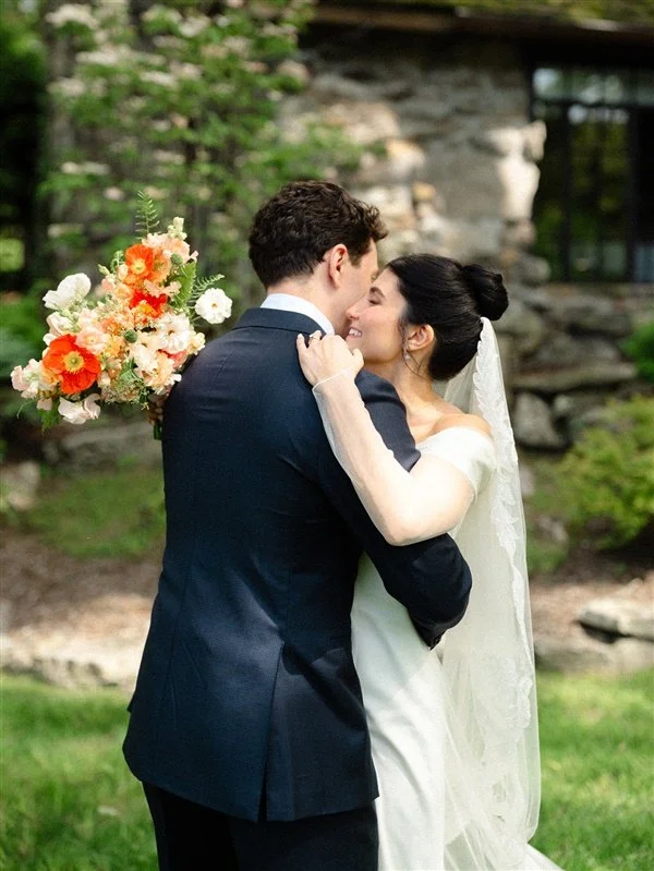 A bride and groom sharing a romantic moment outdoors; the bride is holding a colorful bouquet and smiling as they embrace.