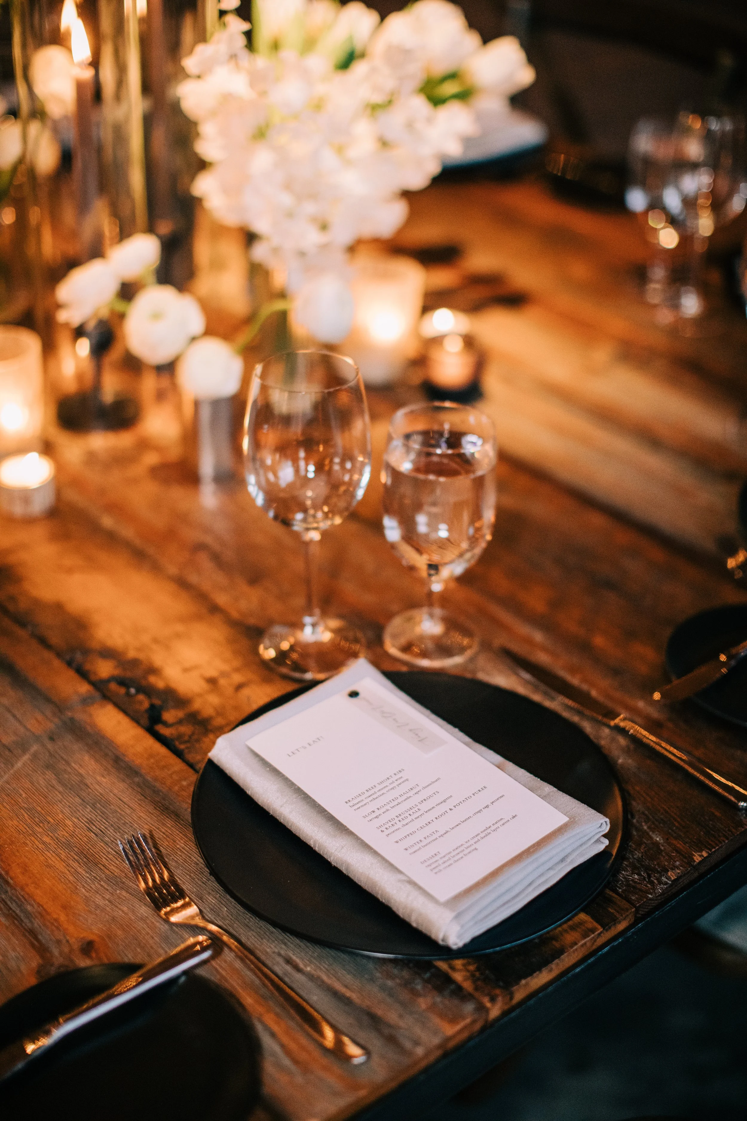 Elegant dining table setting with black plate, white napkin, menu, wine glass, water glass, and floral centerpiece with candles.