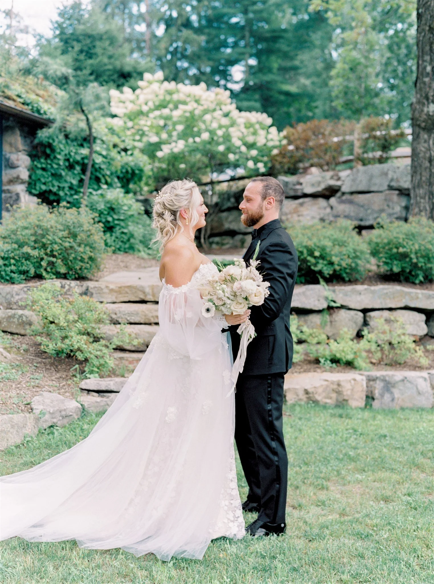 A bride and groom stand outdoors on grass, facing each other, with the bride holding a bouquet of white and blush flowers. The bride wears a white off-the-shoulder wedding gown with lace details, and the groom is dressed in a black tuxedo. They are s