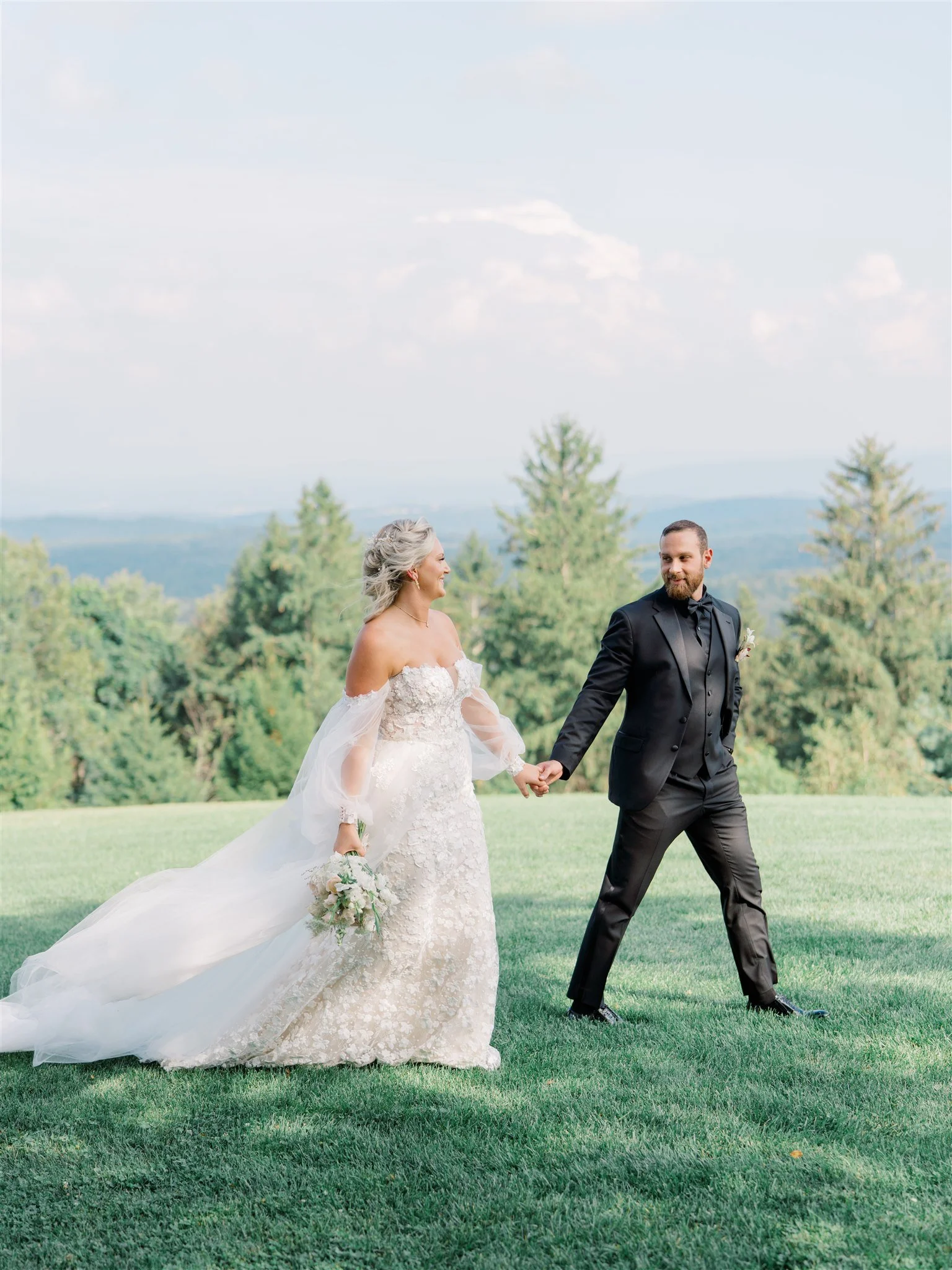 A bride and groom holding hands outdoors on a grassy hill, with trees and a cloudy sky in the background. The bride is in a white wedding dress and the groom is in a black tuxedo.