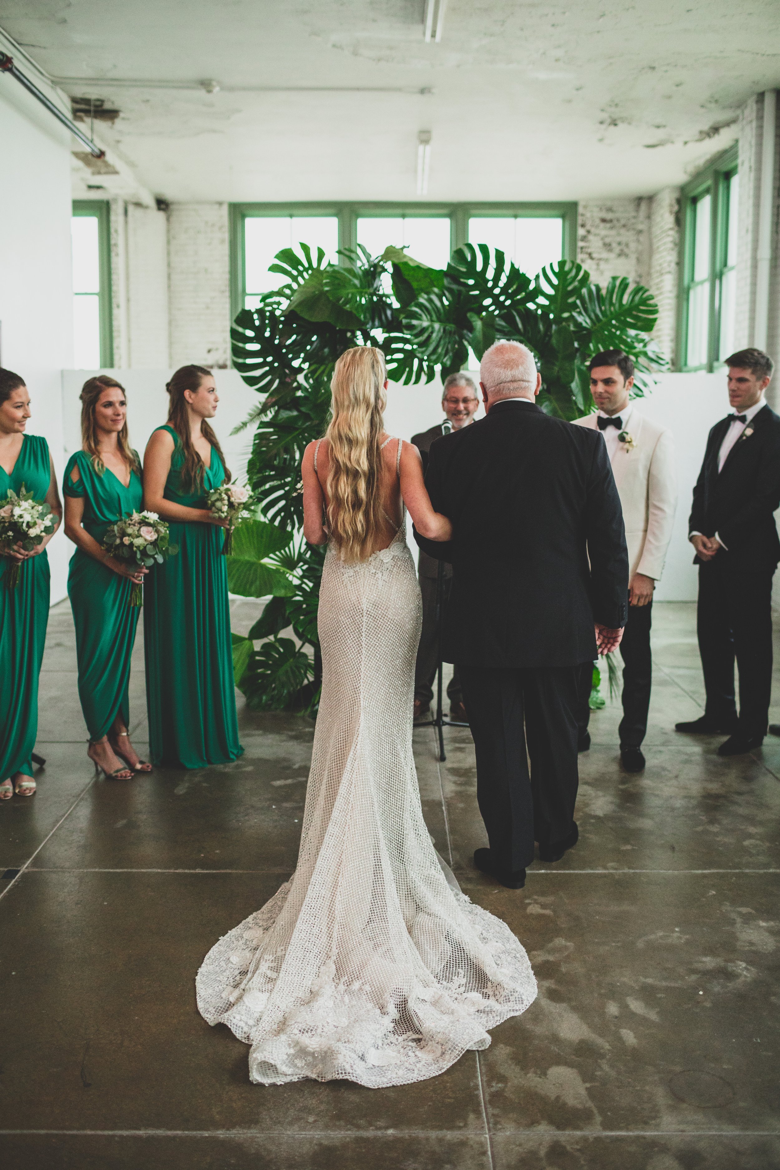 Wedding ceremony with bride in a lace mermaid gown, bridegroom in a white tuxedo, officiant, and bridesmaids in emerald green dresses, inside a studio with large windows and green plants.