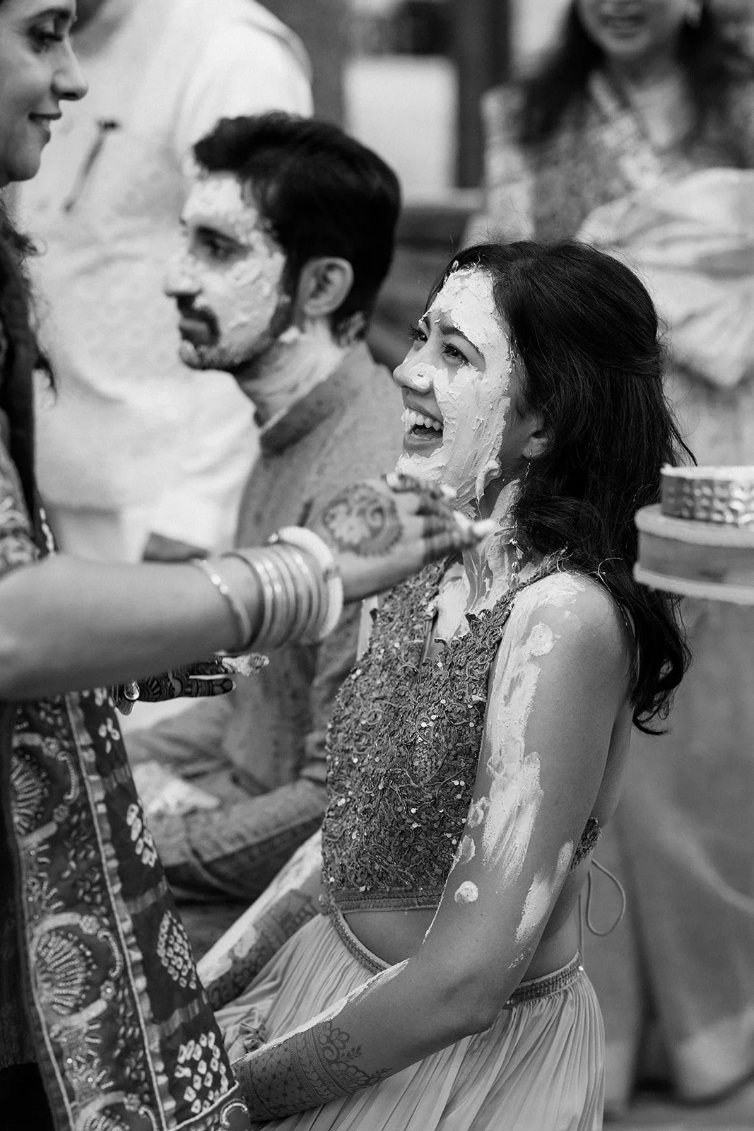 A woman with dark hair and a decorative dress is smiling and being covered in cream by a woman with henna tattoos and bangles during a celebration or ceremony.