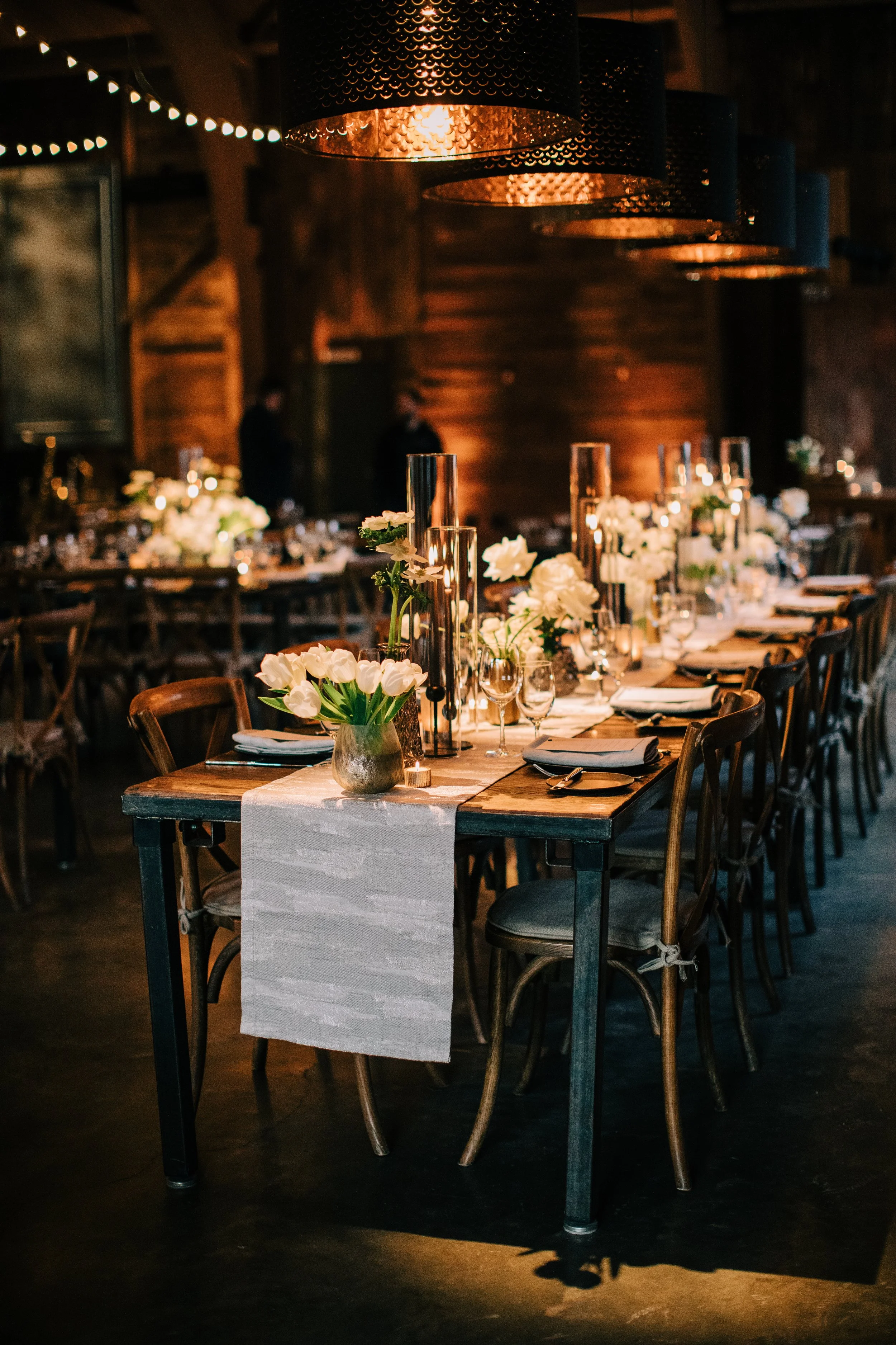 Elegant dining table set for a celebration with white floral arrangements, candles, and black and white tableware in a warmly lit rustic venue.