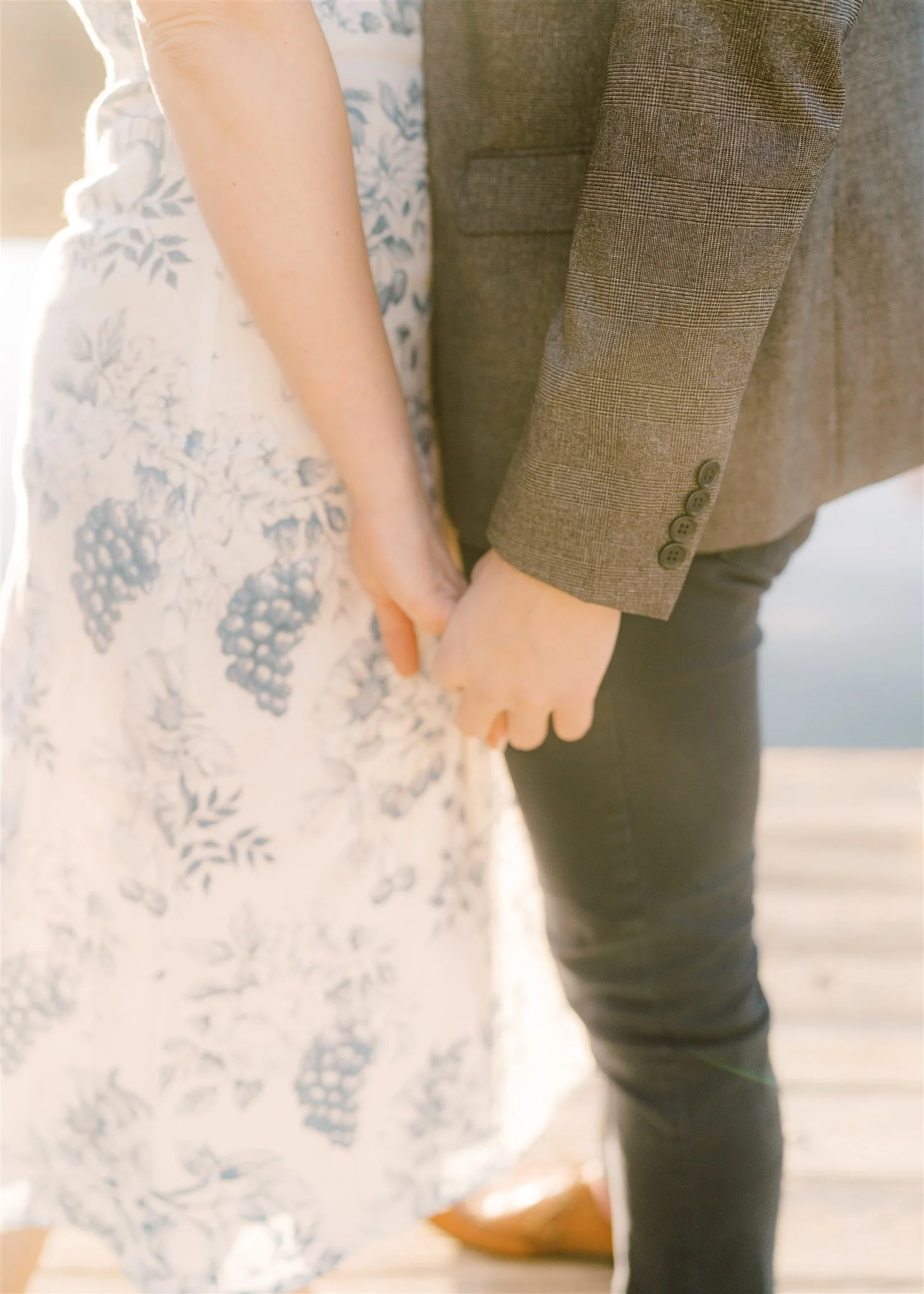 Close-up of a couple holding hands, a woman in a floral dress and a man in a brown suit jacket, outdoors on a wooden deck.